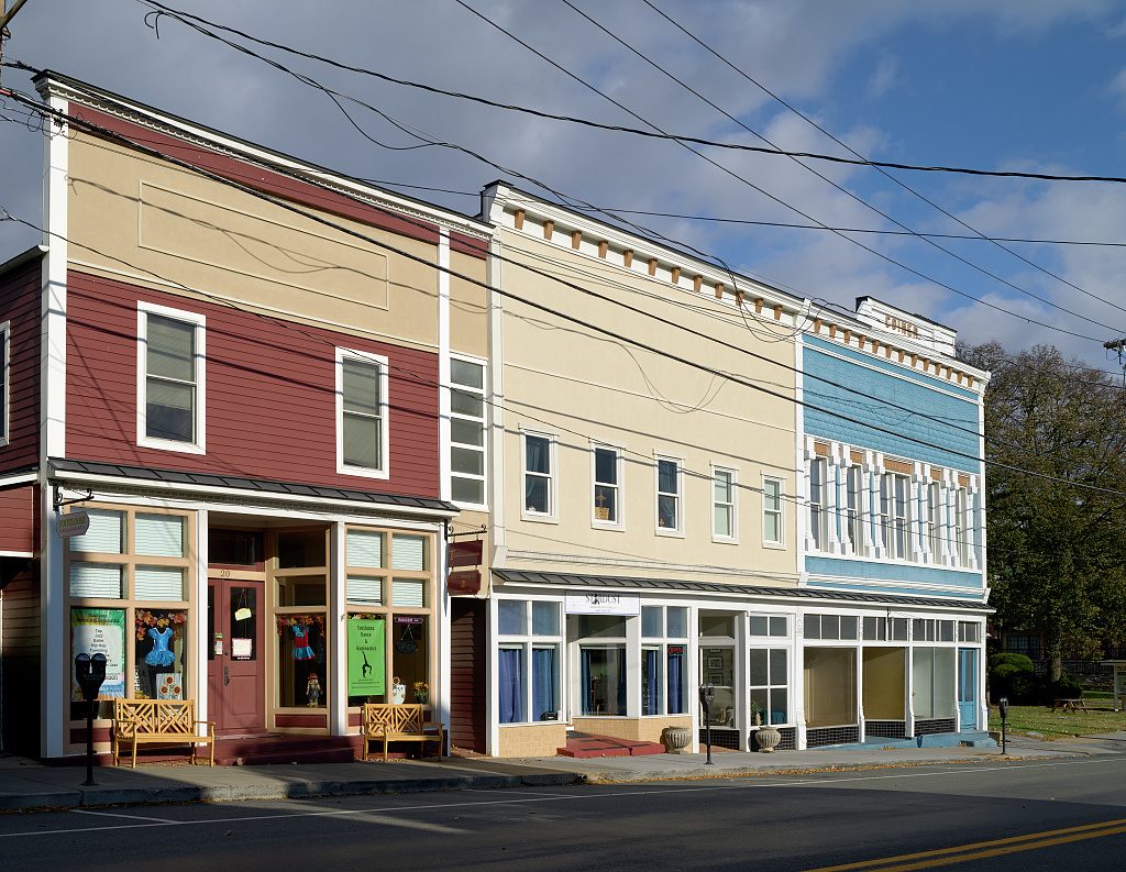 A row of three two-story commercial buildings along a street in late-afternoon sun, with overhead power lines crossing a blue sky. The building at left has a tan upper facade above maroon clapboard, and its shop windows hold costumes and a green poster, with two wooden benches on the sidewalk in front. The middle building is cream-colored with a bracketed cornice and blue curtains in its shop windows, and the building at right has a pale blue upper story with white trim. Parking meters stand along the curb, a double yellow line runs along the road at lower right, and trees and a picnic table on a lawn are at the far right.