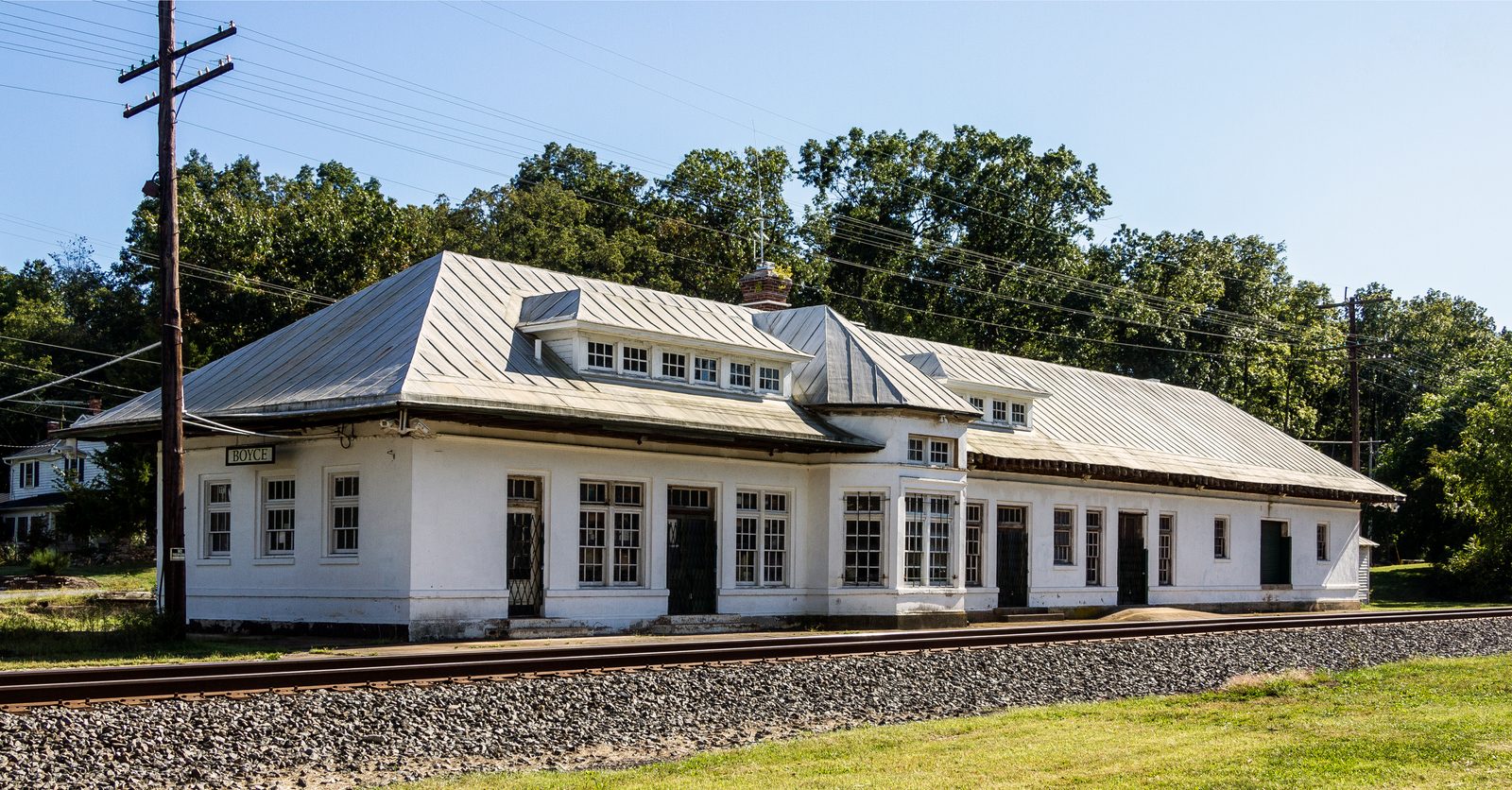 A long one-story white railway station beside a track under a clear blue sky. Its standing-seam metal roof is hipped, with deep eaves, a long dormer holding a row of small windows, a smaller dormer further along and a brick chimney. A projecting bay breaks the front wall, and multi-pane windows alternate with dark doors, some behind folding metal gates. A small sign reading BOYCE hangs under the eaves at the left end. Gravel ballast and rails run across the foreground, wooden utility poles carry wires at left and right, and tall trees in full leaf rise behind the building.