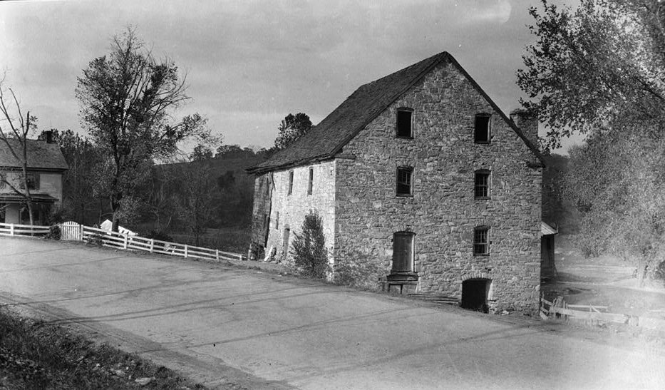 A black-and-white photograph of a tall stone mill beside a curving dirt road. The mill has three stories and an attic under a steep roof, rows of small square windows in its gable end, a raised doorway with a small wooden stoop, and a low dark arched opening at the base. Its long side wall, turned toward the left, is paler than the gable, a chimney rises at the far end of the roof, and a low shed stands against the right side. At left, a white board fence with a gate runs along the road toward a two-story frame house with a porch, and trees stand in front of a wooded hill.
