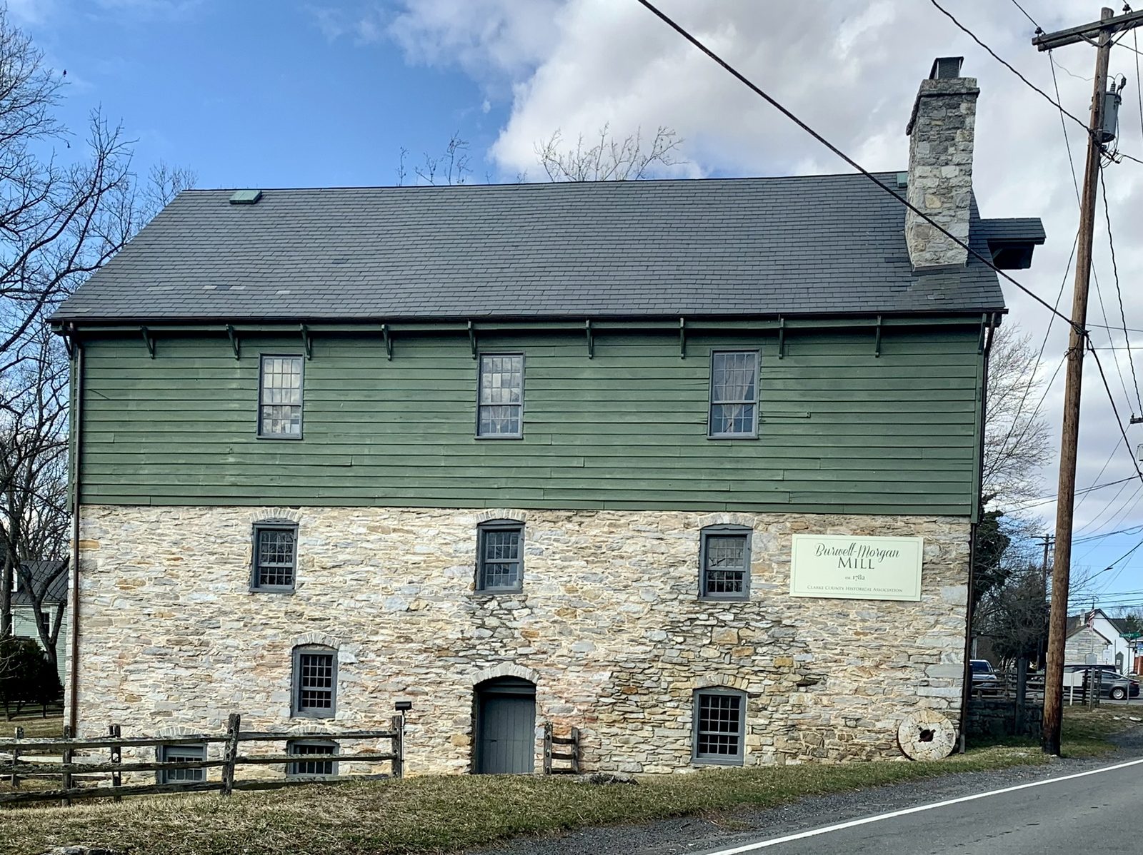 A two-storey mill building stands close to a road under a blue sky with broken cloud. Its lower storey is coursed rubble limestone pierced by small barred windows and a low arched doorway with a plank door; the upper storey is clad in weatherboard painted dark green, with three twelve-pane sash windows. A steep grey shingled roof runs the length of the building and a stone chimney rises at the far gable. A painted board on the stonework reads Burwell-Morgan Mill, est. 1782, Clarke County Historical Association, and a millstone leans against the wall beside it. A split-rail fence crosses the foreground at the left and a utility pole and wires stand at the right.