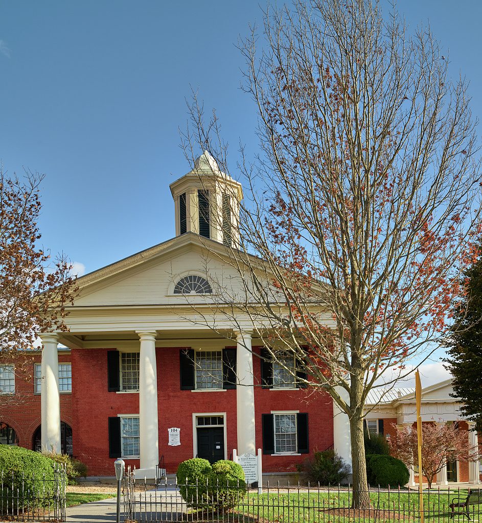 A red brick courthouse of two stories under a clear blue sky, fronted by a white portico of four tall columns that carries a pediment with a half-round fanlight. An octagonal white cupola with louvered openings rises from the roof. The windows have black shutters, a small white plaque hangs beside the central door, and a white sign stands on the lawn. A black iron fence and rounded boxwood shrubs line the front, a bare tree holding a few red leaves stands at right, and a lower building with white trim adjoins at the far right.