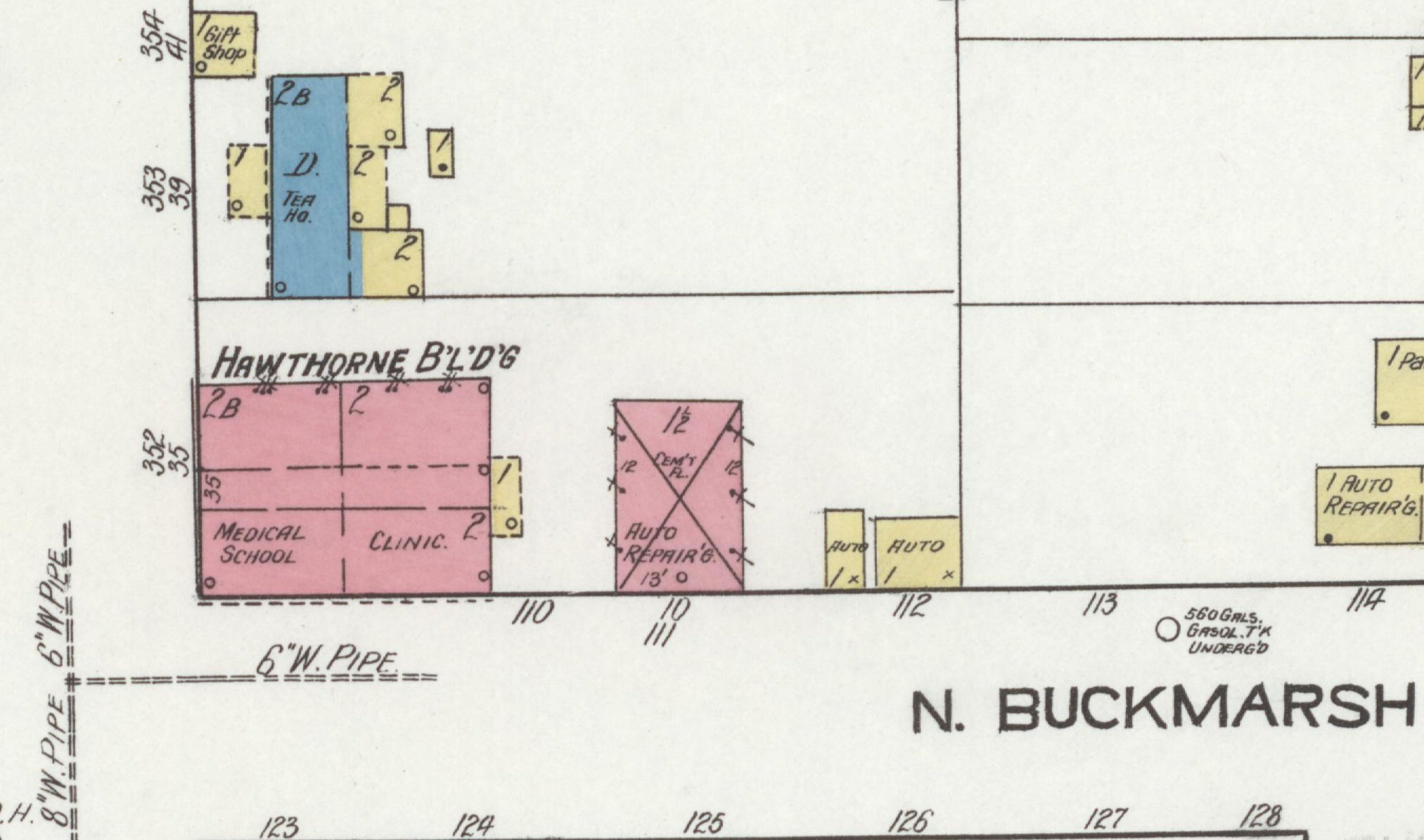 A detail of a hand-colored fire insurance map. At its center is a block tinted pink, the Sanborn color for brick, lettered HAWTHORNE B'L'D'G along its top edge and divided into sections marked 2B, 2, MEDICAL SCHOOL and CLINIC, with the numbers 352 and 35 beside it. To its right are a smaller pink building crossed by an X and marked AUTO REPAIR'G and CEM'T FL., and two small yellow sheds marked AUTO. Above it stand a blue building marked D. and TEA HO., yellow outbuildings and a small yellow Gift Shop. Dashed lines marked 6" W. PIPE and 8" W. PIPE run through the open ground, lot numbers run along the frontages, and the street name N. BUCKMARSH is printed at lower right.