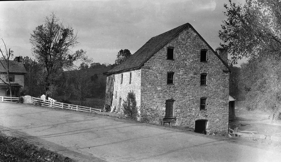 Black-and-white photograph of the Burwell Mill at Millwood, a tall stone building with a steep roof beside a curving road, a white fence and a frame house.