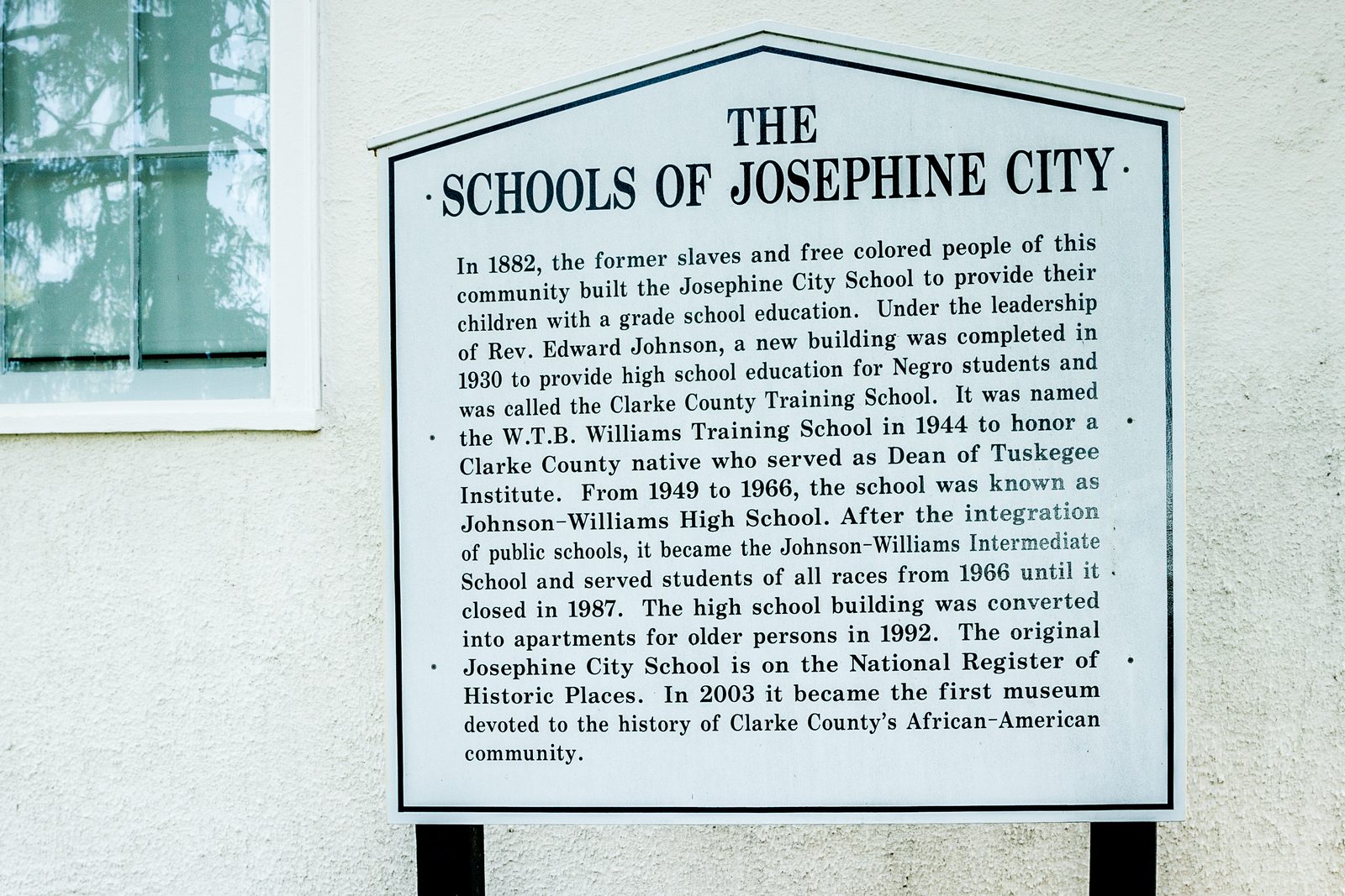 A white sign shaped like the front of a house, with a pointed top and a dark border, stands on two black posts against a textured cream stucco wall. Its heading reads THE SCHOOLS OF JOSEPHINE CITY in bold capital letters, and a paragraph of smaller type about the schools' history fills the rest of the sign. At upper left, a multi-pane window reflects the branches of a tree.