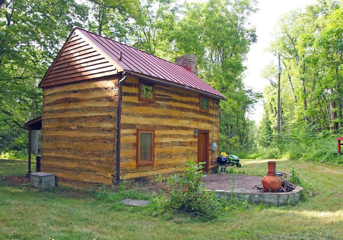 The cabin from behind in summer: a gable end of hewn logs under a faded red metal roof, a door with a small porch roof, and a stone-edged patio with a clay chiminea on the grass beside it.