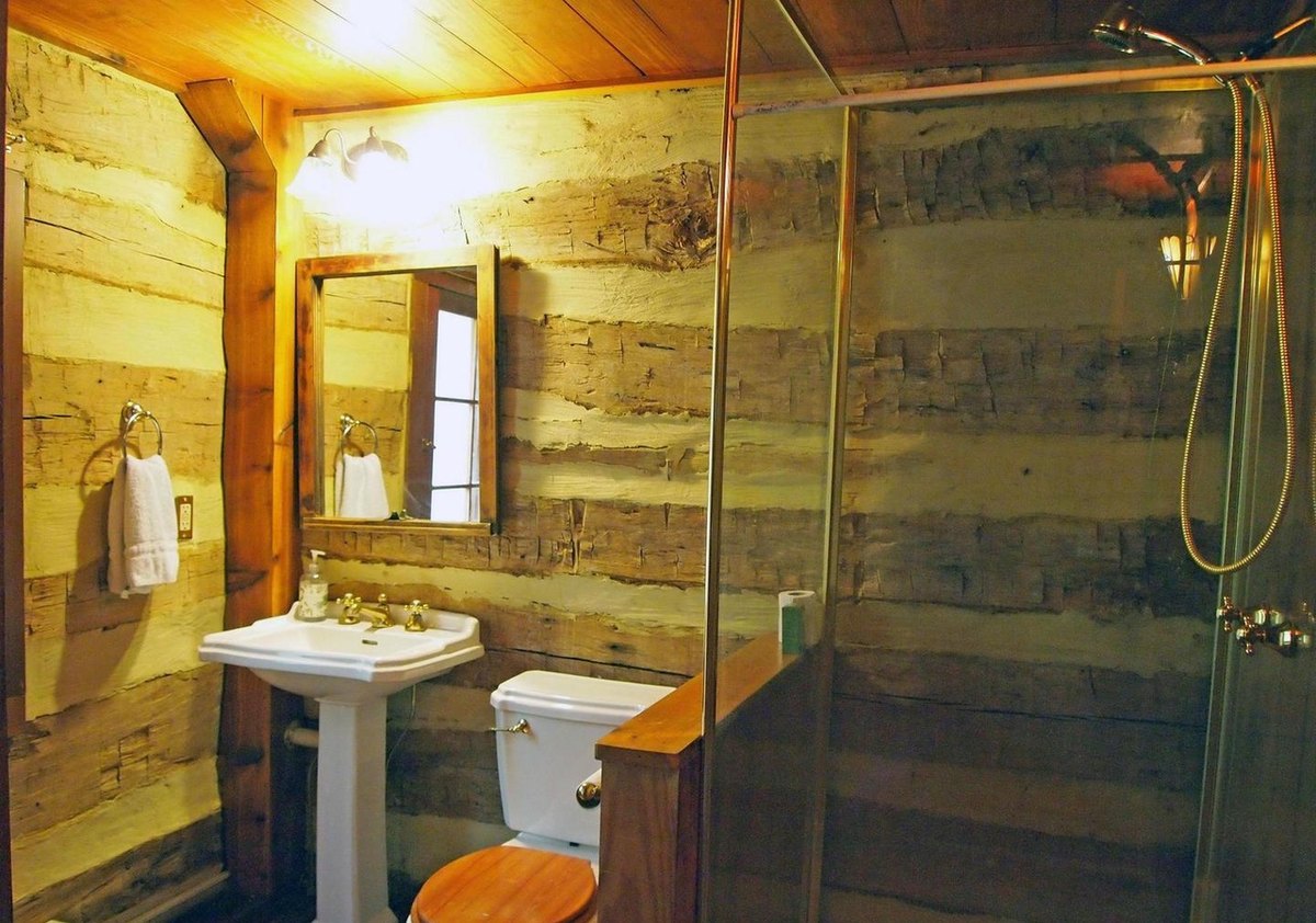 The bathroom with log walls, a pedestal sink under a mirror, a wooden toilet seat and a glass shower enclosure.