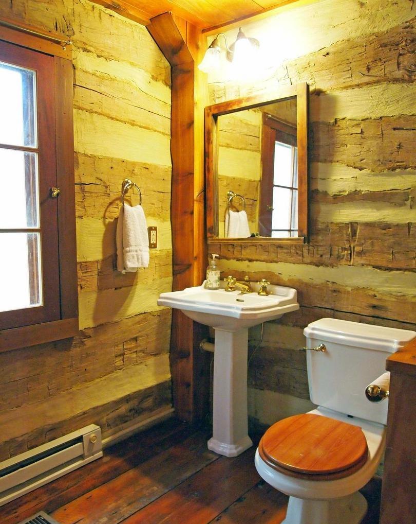 The bathroom from another angle: a window in the log wall, a towel ring, a pedestal sink and mirror, and a toilet with a wooden seat.