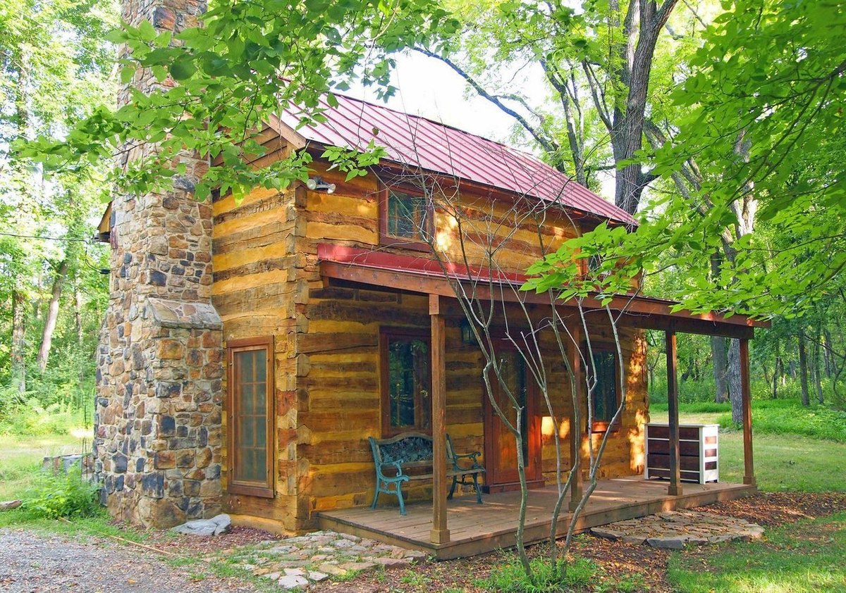 The cabin at an angle showing the stone chimney end and the long covered front porch with a bench and green chairs, partly screened by a young tree.