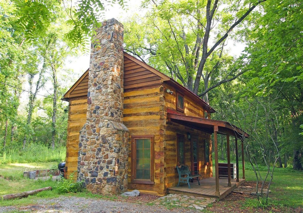 The chimney end of the cabin in summer: a tall fieldstone chimney on the log gable, a narrow glazed door beside it and the covered front porch turning the corner, among tall trees.