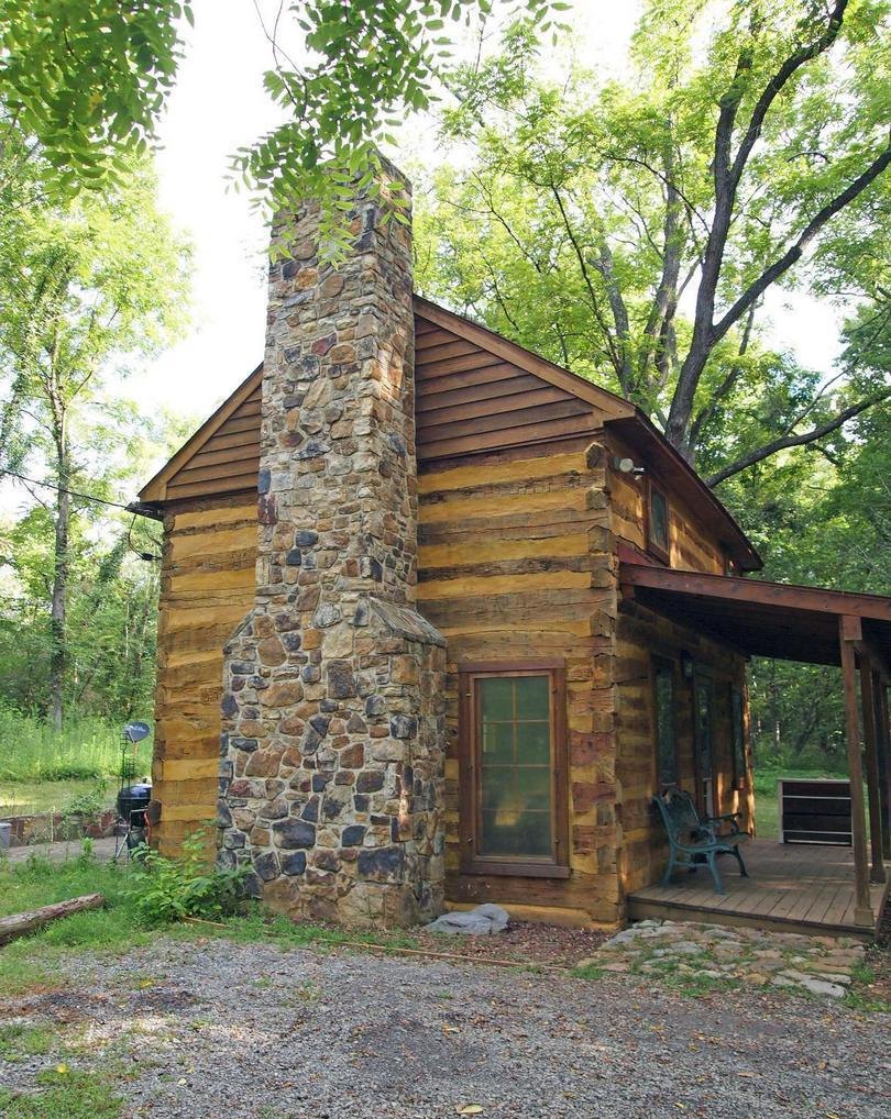 A closer upright view of the fieldstone chimney rising against the log gable end, with the glazed door at its side and green chairs on the porch beyond.