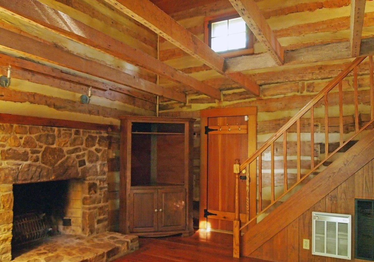 The main room showing the corner of the stone fireplace, a wooden corner cabinet, a board door and the stair climbing along the wall, under ceiling beams and a small gable window.