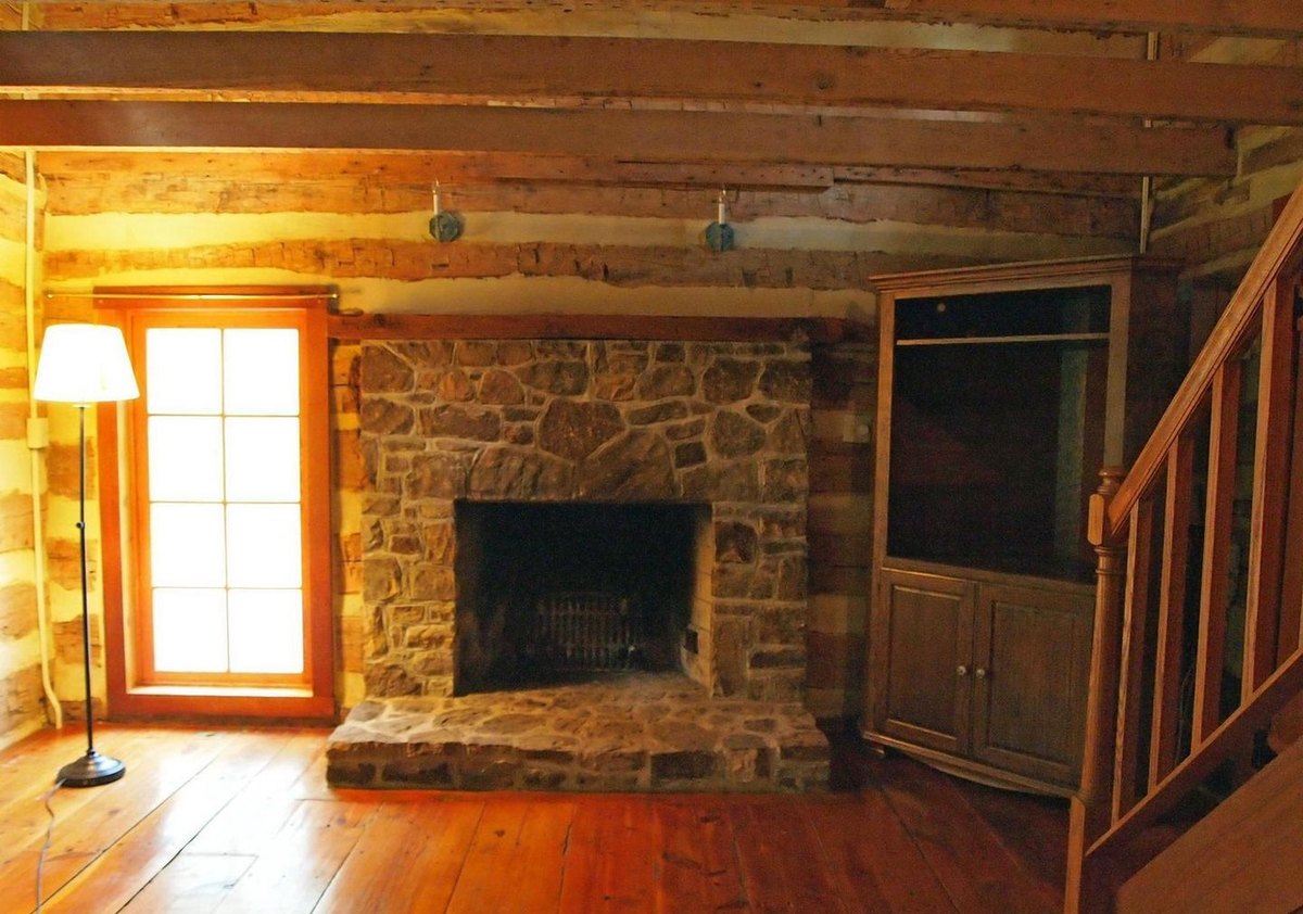 The stone fireplace with an open hearth and no stove, between a glazed door and a corner cabinet, with the stair rail at the right and wide floorboards.