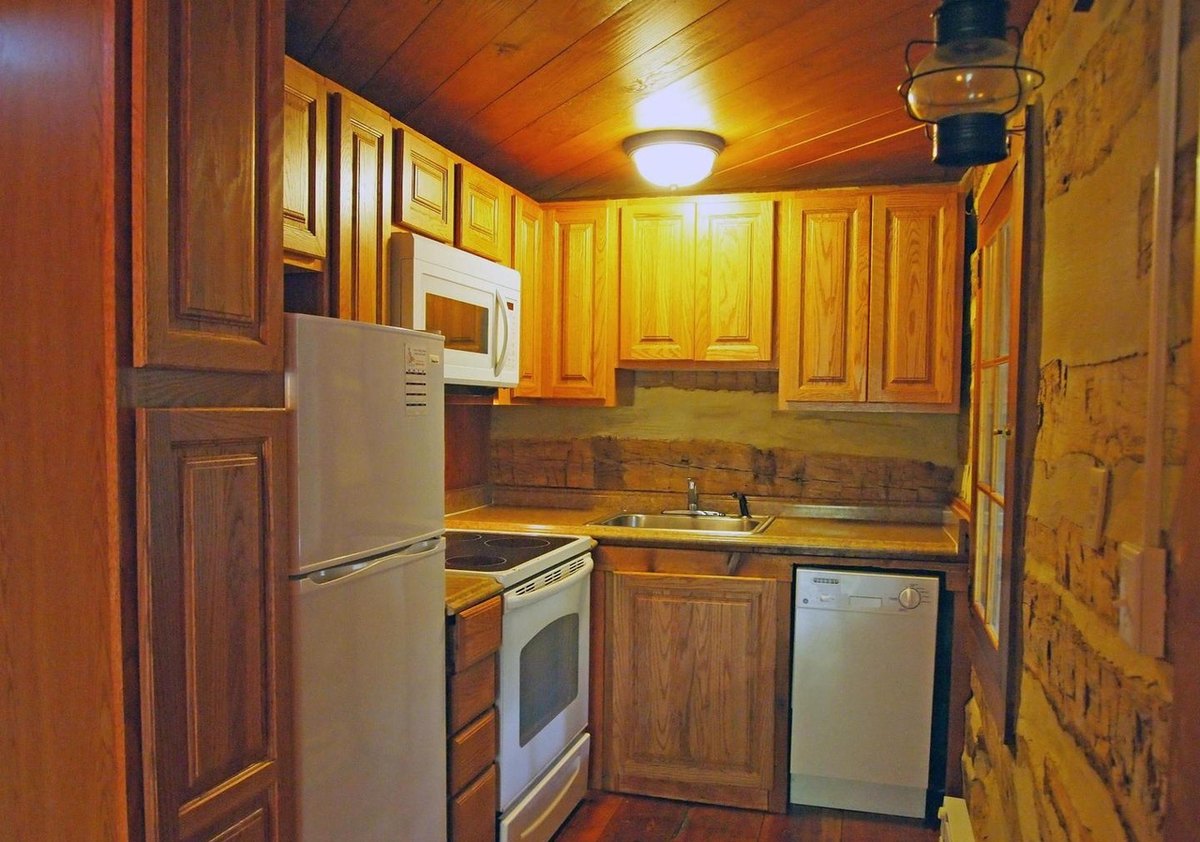 The small kitchen with wooden cabinets, a white refrigerator, stove, microwave and dishwasher, a board ceiling with a round light, and log walls at the side.