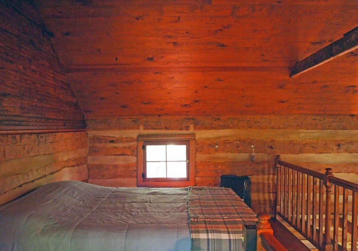 The upstairs room under a sloping board ceiling, with a bed, a small window in the log gable and a wooden stair railing.