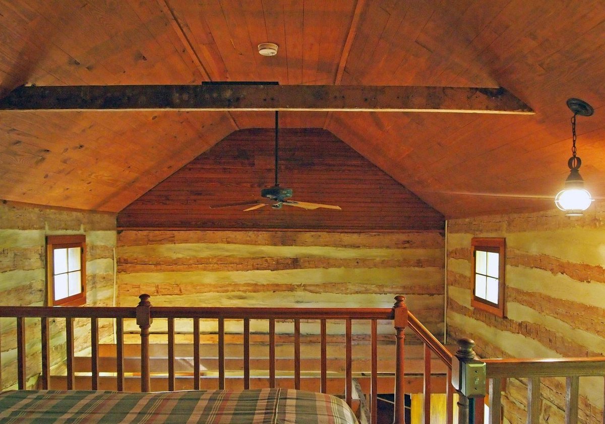 The upstairs room toward its gable end: a board ceiling with a ceiling fan and hanging lantern, two small windows in the log walls, and the stair railing across the foreground.