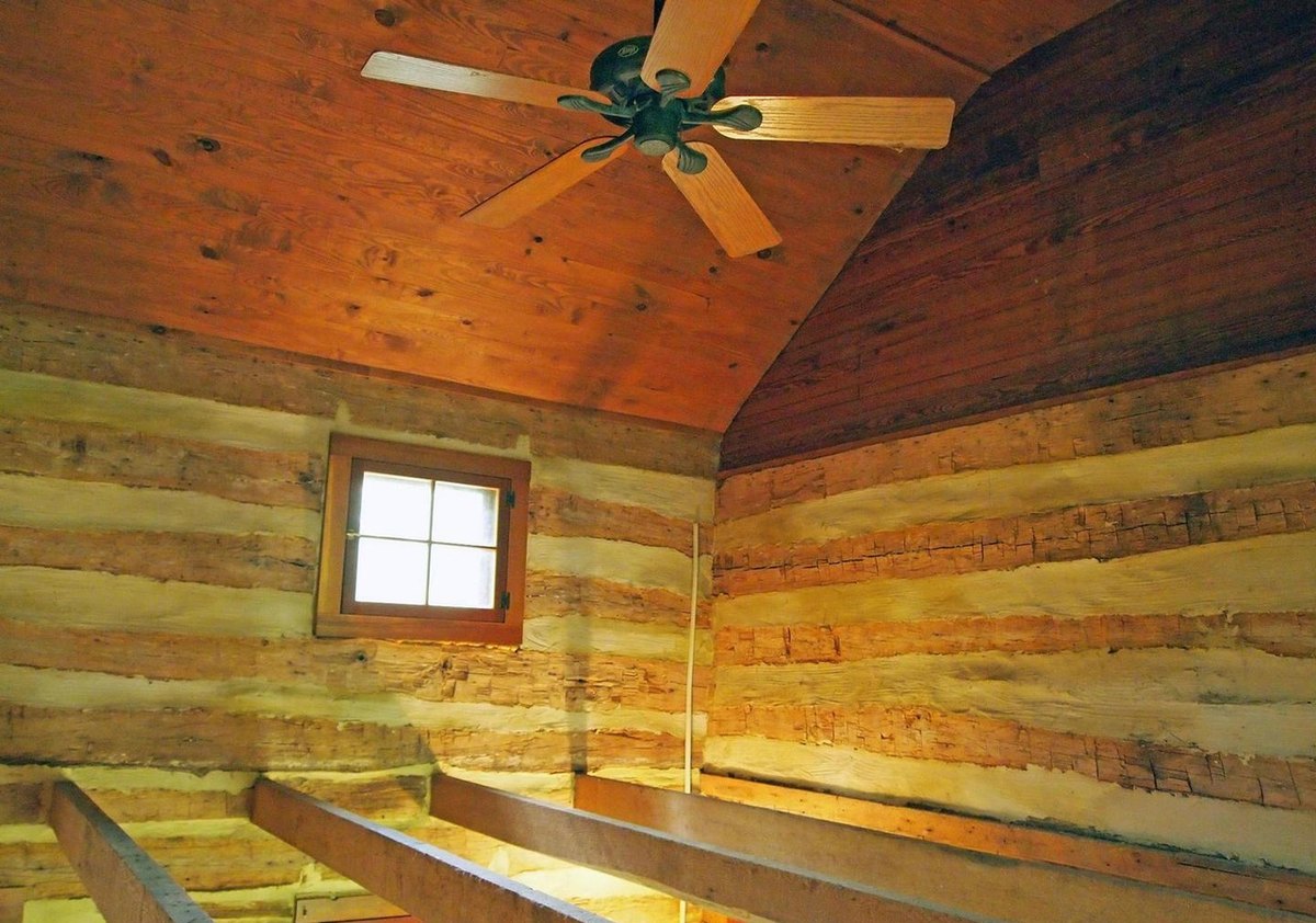 A small square window in a log gable wall below a sloping board ceiling with a ceiling fan, and low beams across the floor.