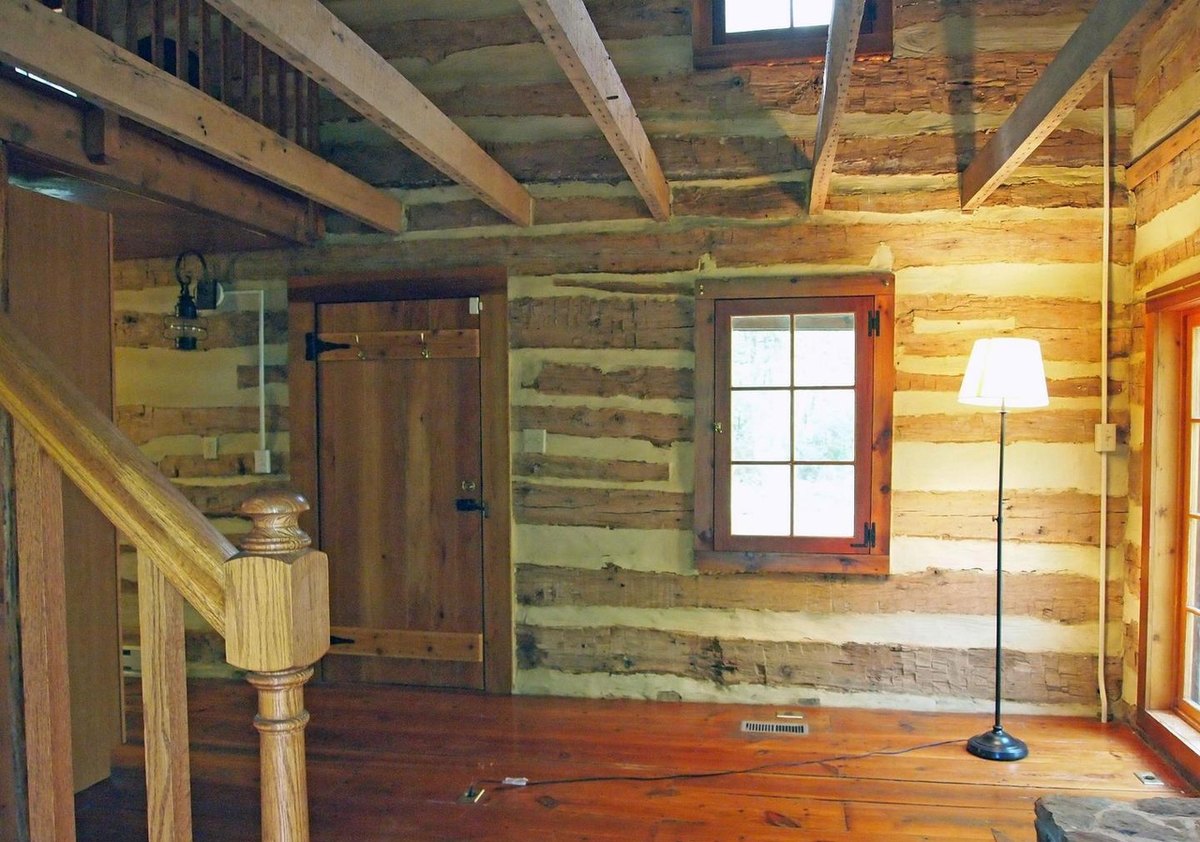 The empty main room: log walls with pale chinking, exposed ceiling joists, a board door, a small window, a floor lamp, and the newel post of the stair in the foreground.