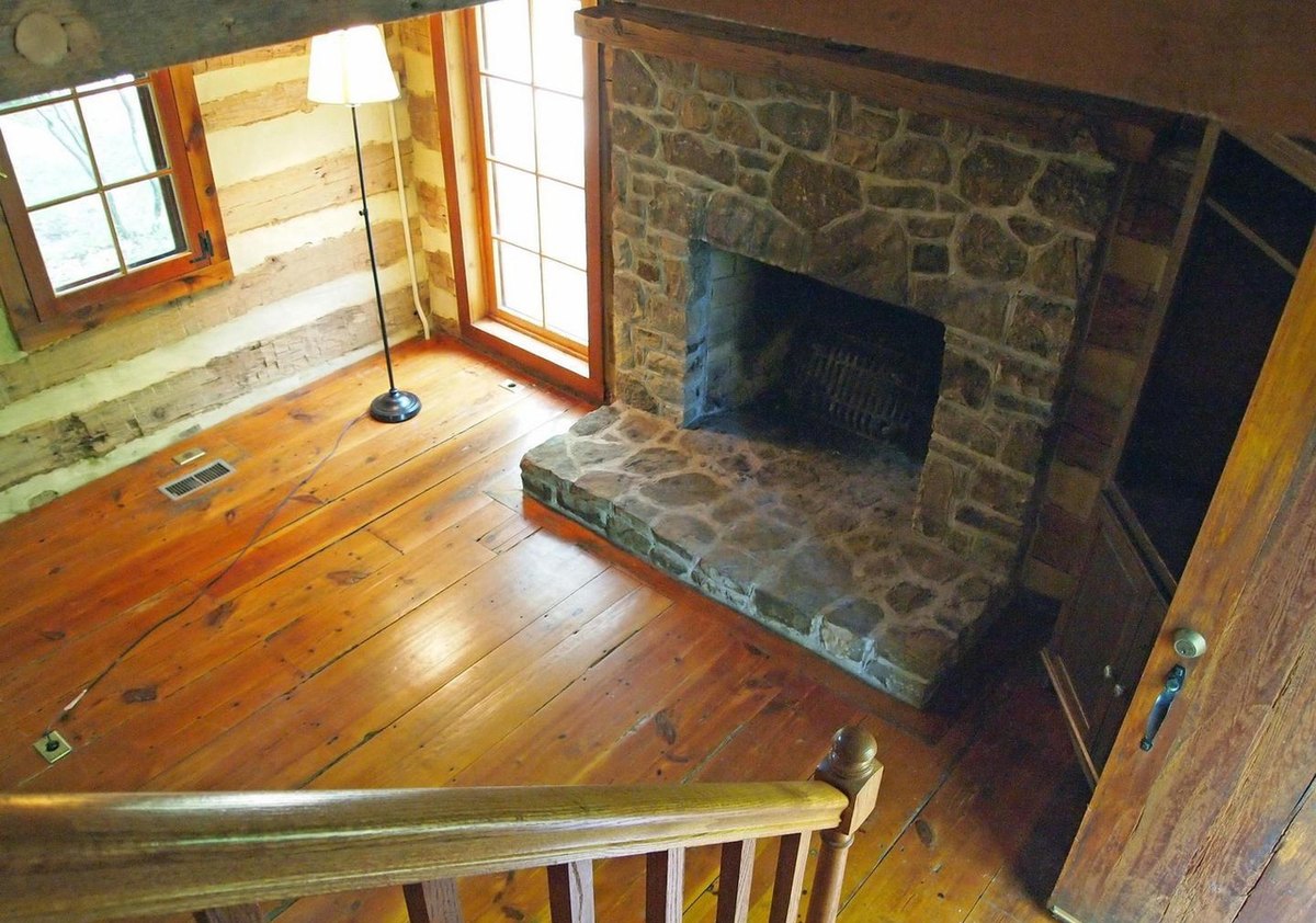 Looking down from the top of the stair onto the main room's plank floor and the stone fireplace, with a floor lamp and windows beside it.