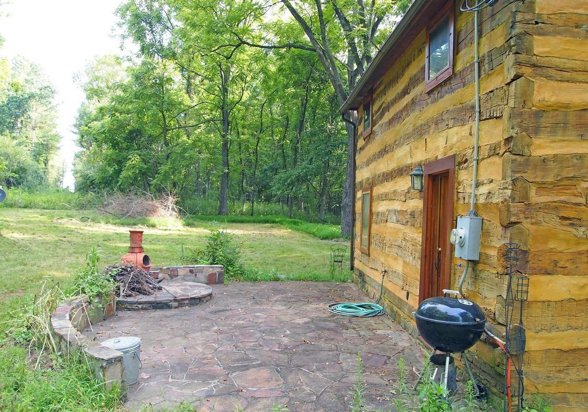 A flagstone patio along the log wall of the cabin, with a kettle grill, a door and electric meter on the wall, and a low curved stone wall with a chiminea beyond.