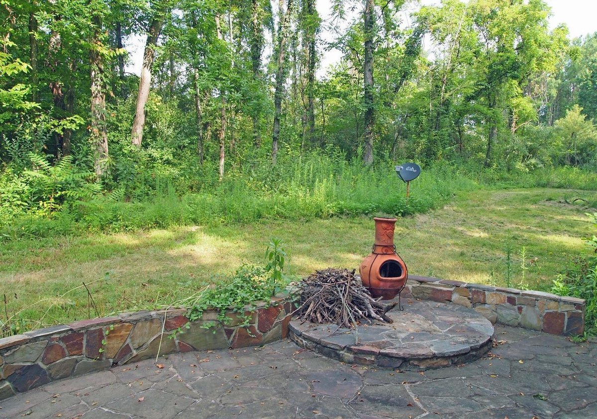 The round flagstone patio behind the cabin, edged by a low curved stone wall, with a clay chiminea and a pile of branches on a raised stone circle and woods behind.