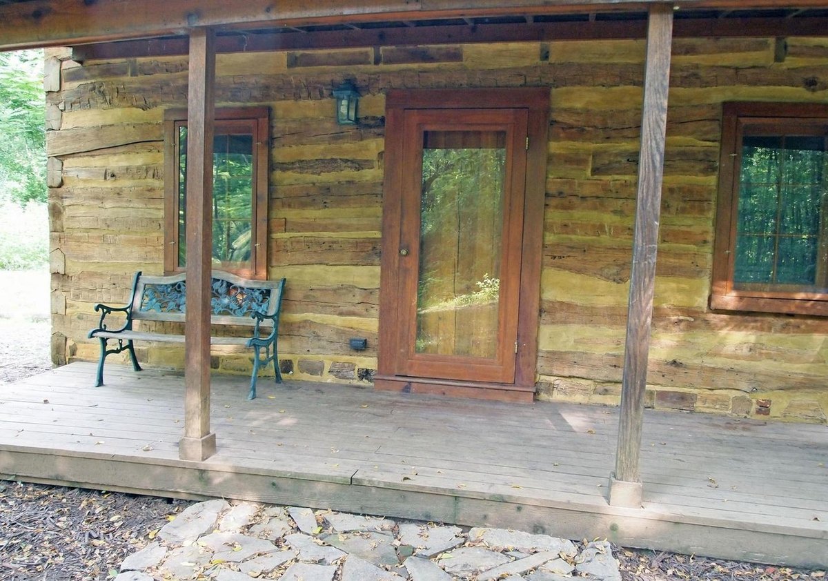 The front porch close up: a wooden front door between two windows in the log wall, an iron and wood bench, and a flagstone step in front of the porch floor.
