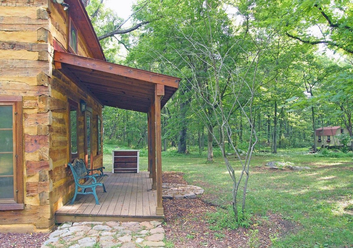 The front porch seen from its end: a blue bench against the log wall, the porch roof on wooden posts, and a small red-roofed shed far off among the trees.