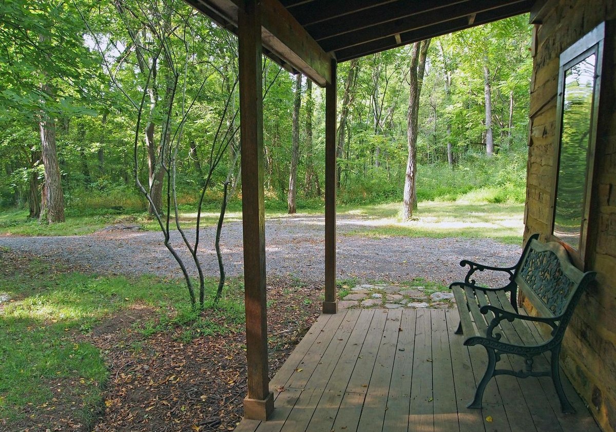 The view from the front porch past a bench and the porch posts to the gravel drive and the woods beyond.