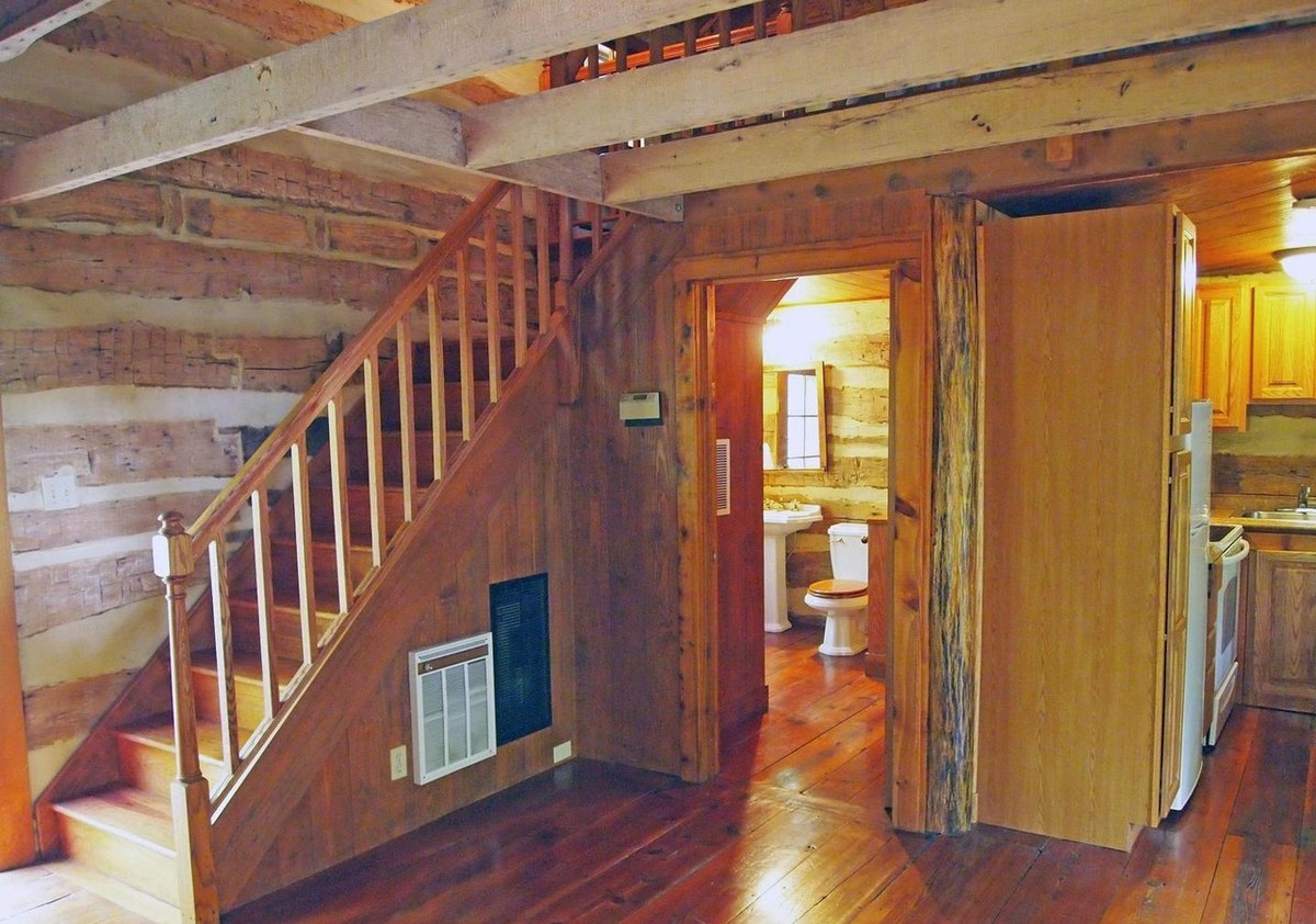 The wooden stair rising along a log wall, with an open door to the bathroom beside it and a glimpse of the kitchen through a second doorway.