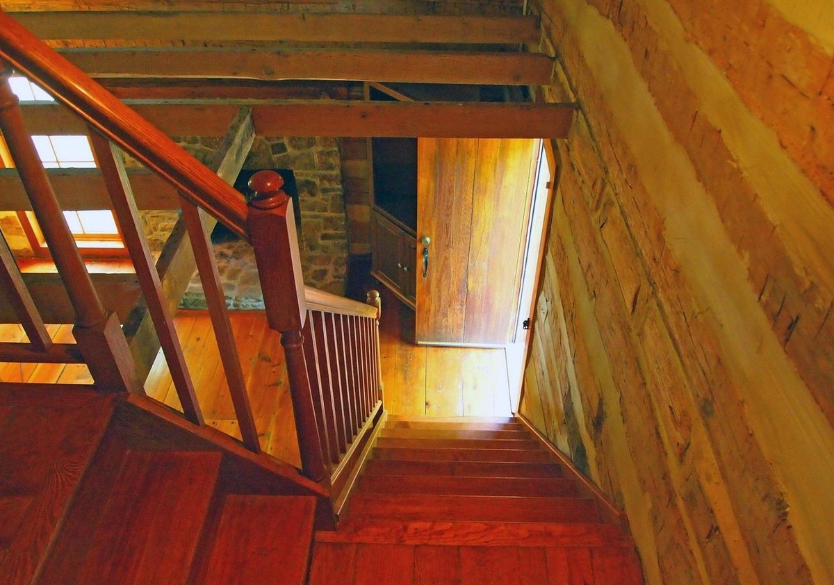 Looking down the narrow wooden stair from the upper floor, with its railing, log walls and the lit room below.