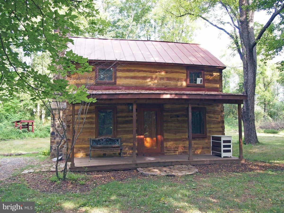 A two-story cabin of squared, hewn logs with pale chinking and a faded red metal roof, seen from the front under leafy trees. A covered porch runs across it with a wrought-iron bench and a small chest of drawers beside the door, and a fieldstone chimney rises at the left end.