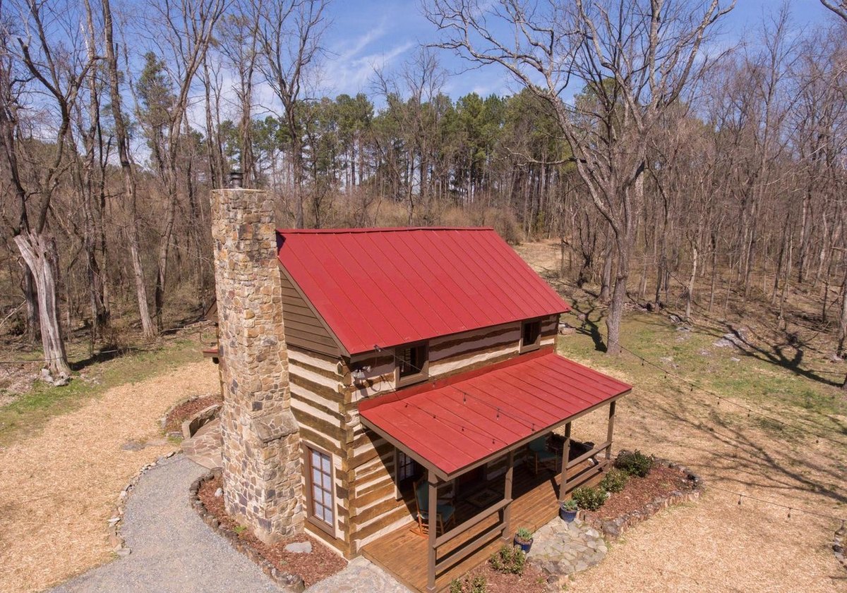 A low aerial view of the cabin from the chimney-end corner, showing its red roof, the stone chimney, the front porch and the gravel path, in bare early-spring woods.