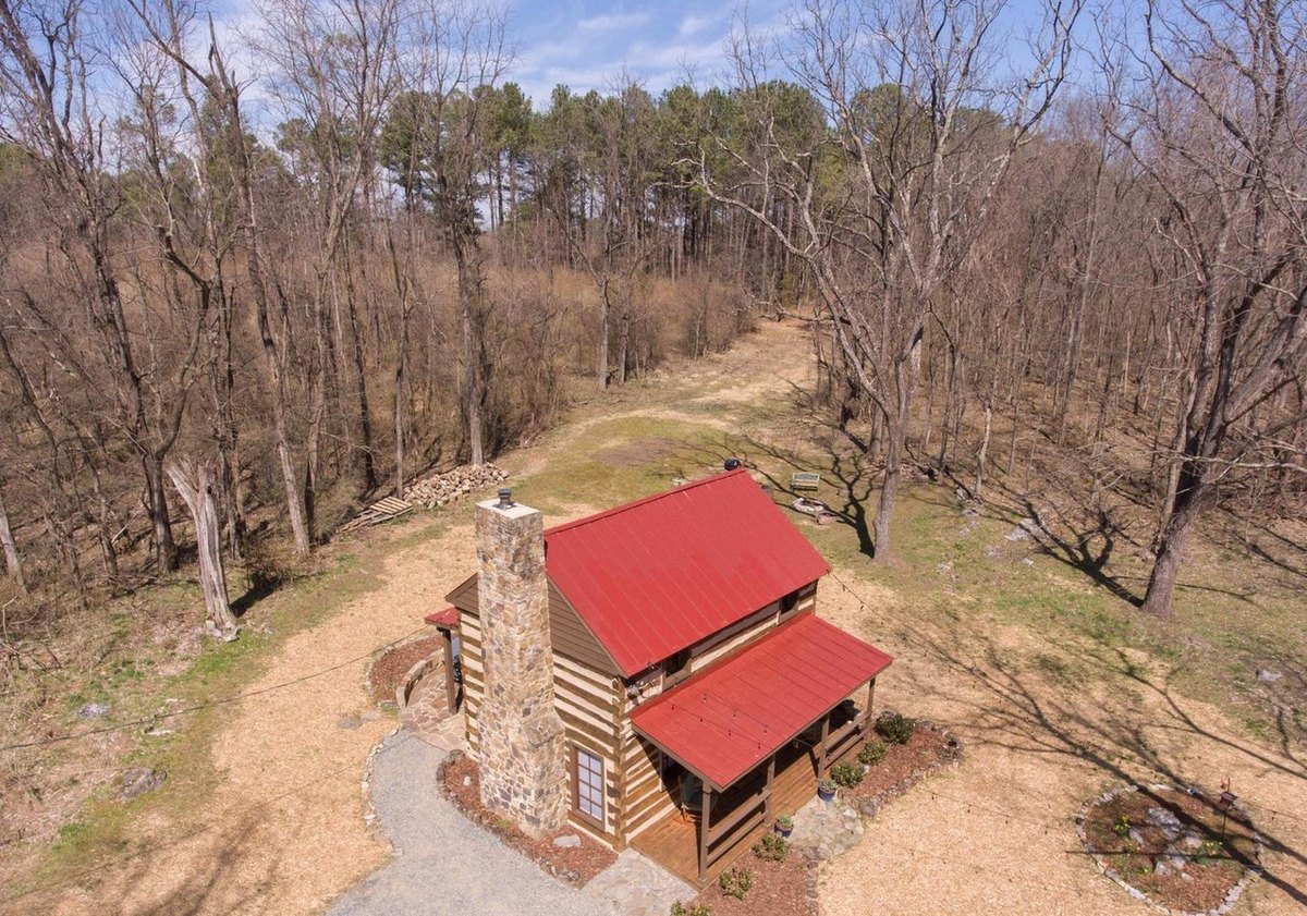 An aerial view of the cabin from its chimney side, with the red roof, the chimney, a curving gravel path and mulched clearing, and a track leading off into the woods.