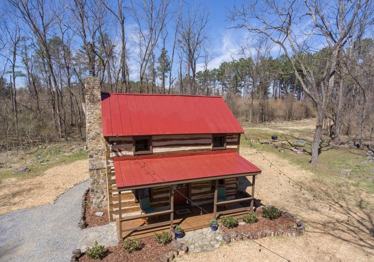 An aerial view of the front of the cabin: the red roof and porch roof, the stone chimney at the left end, the porch with planters, and the gravel drive.