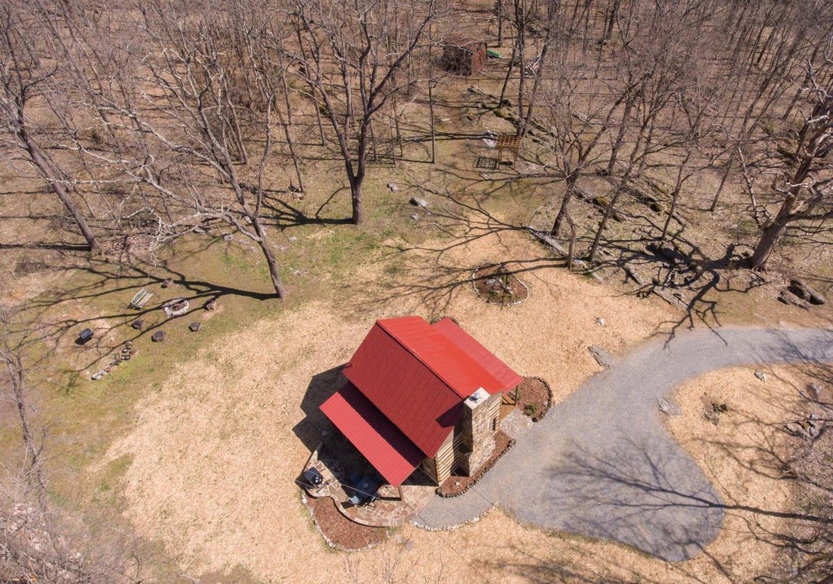 Looking almost straight down on the cabin's red roof and chimney in a mulched clearing, with the gravel drive curving away between bare trees.