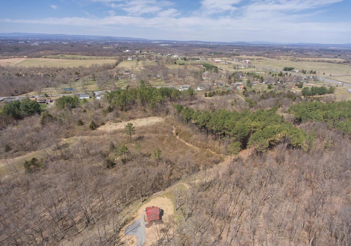 A high aerial view over bare woods and open farmland toward distant mountains, with the small red-roofed cabin in a clearing at the lower edge.