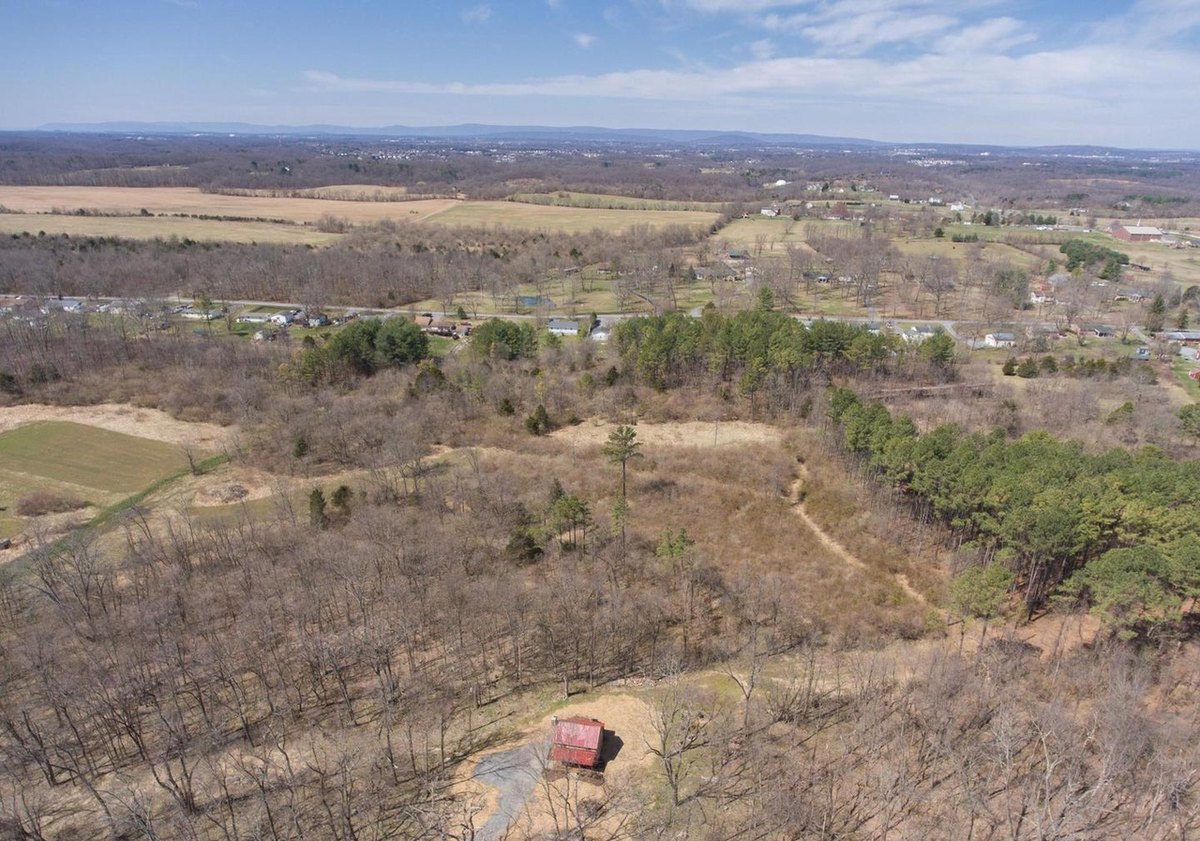 An aerial view across woods, fields and scattered houses to a far mountain ridge, with the cabin and its drive small in the foreground.