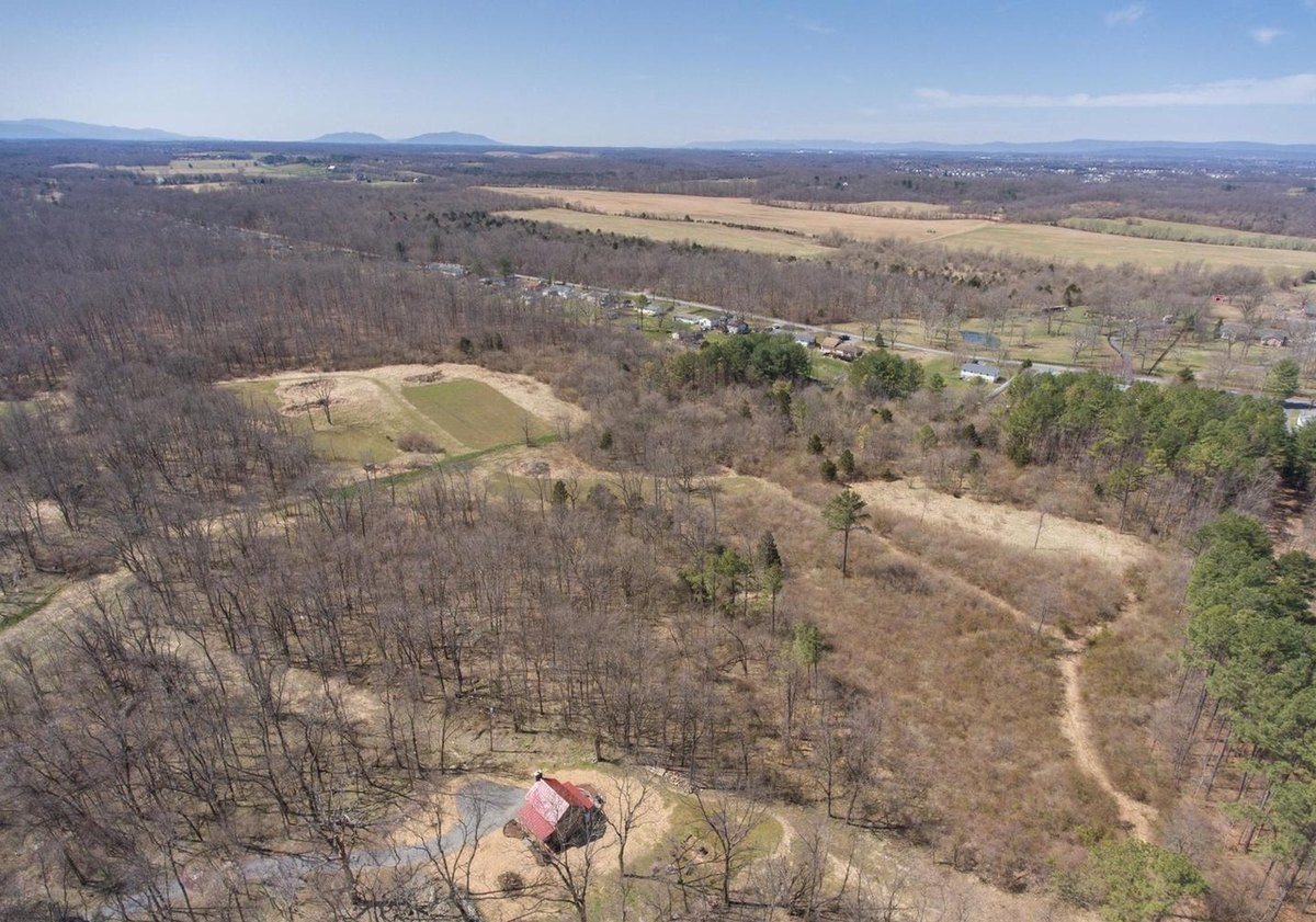 An aerial view of the cabin in its clearing among bare trees, with tracks through the woods and open fields and a mountain ridge on the horizon.