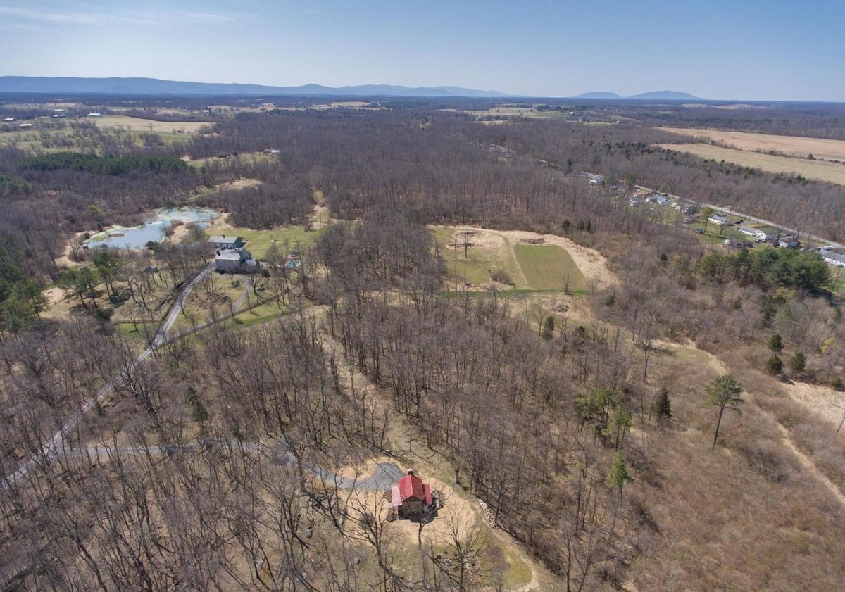 An aerial view with the cabin in the foreground, a drive winding through bare woods, a pond and houses in the middle distance and mountains beyond.