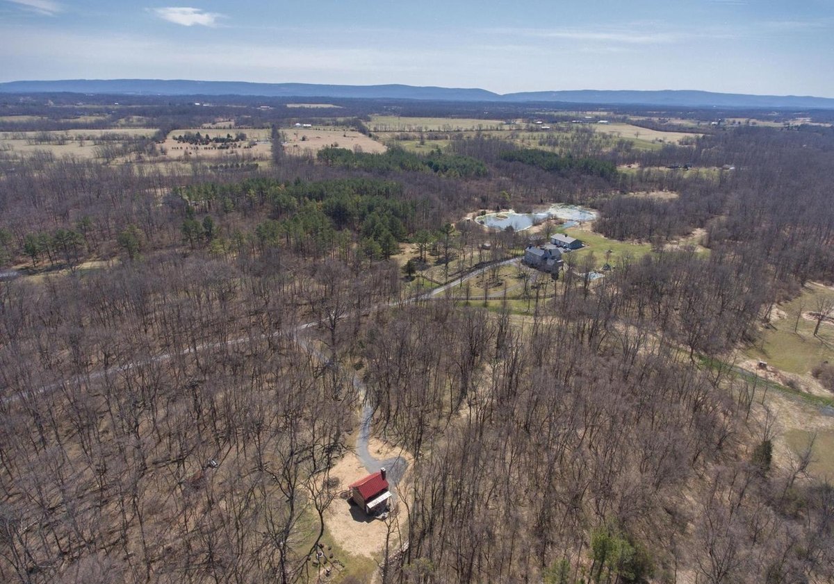 The cabin in a mulched clearing seen from above, with its drive leading through bare woods toward a house and pond and fields reaching to the mountains.