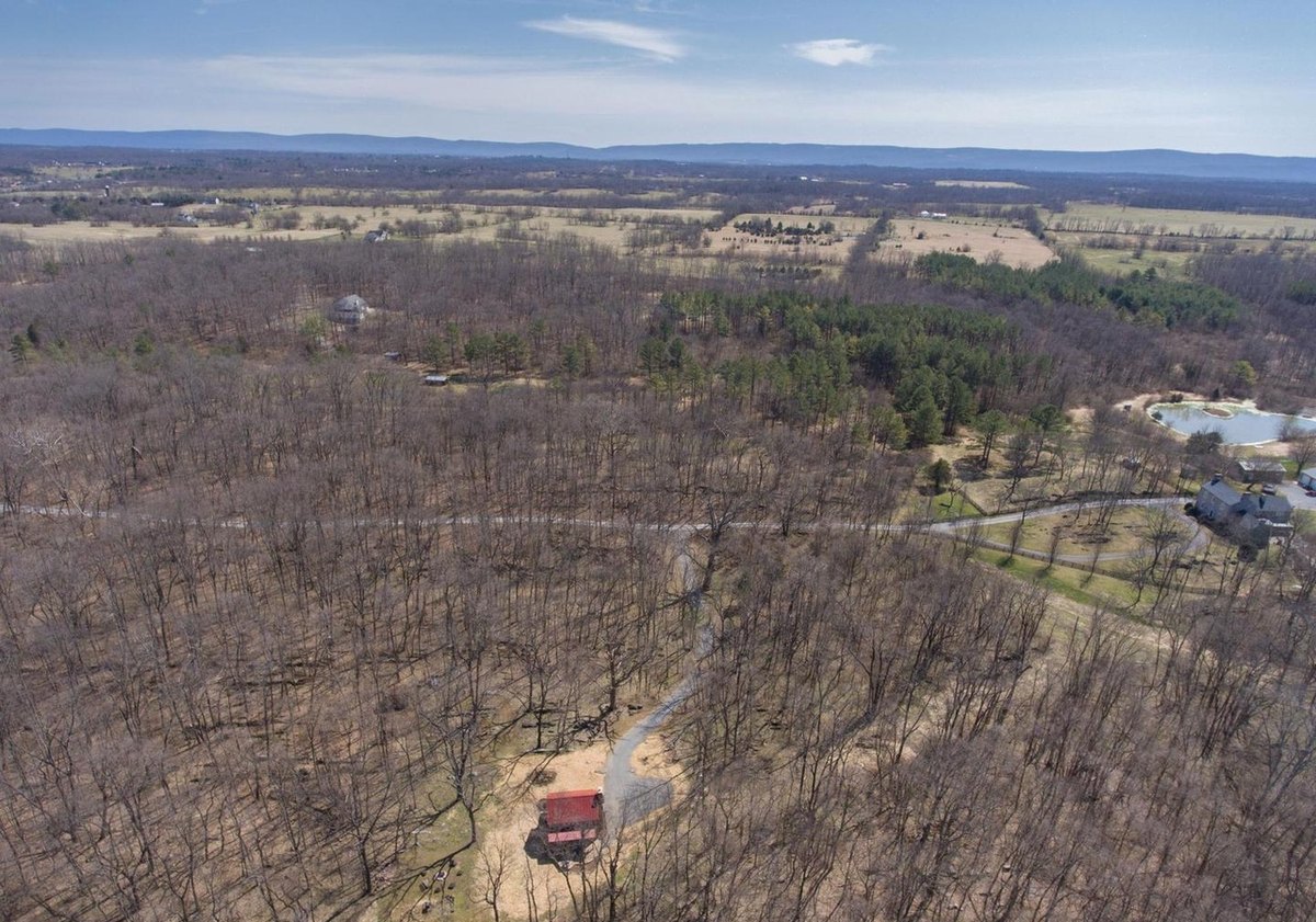 An aerial view of the gravel drive curving through bare woods to the small red-roofed cabin, with houses, fields and mountains in the distance.
