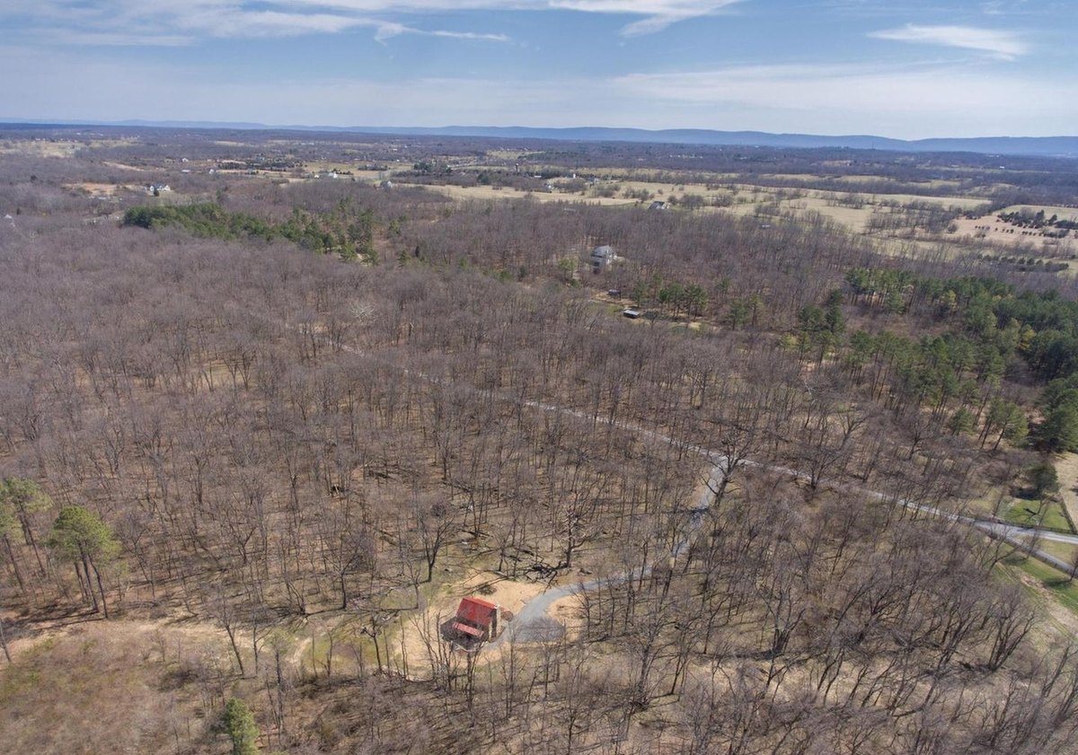 A wide aerial view over bare woods with the small red-roofed cabin in a clearing near the bottom and farmland and mountains on the horizon.