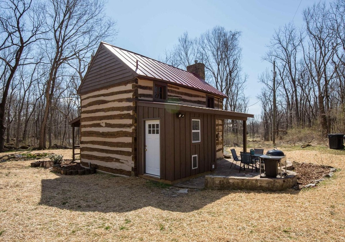 The back of the cabin: a board-sided addition with a white door against the log walls, the patio with a table and chairs, and bare trees around.