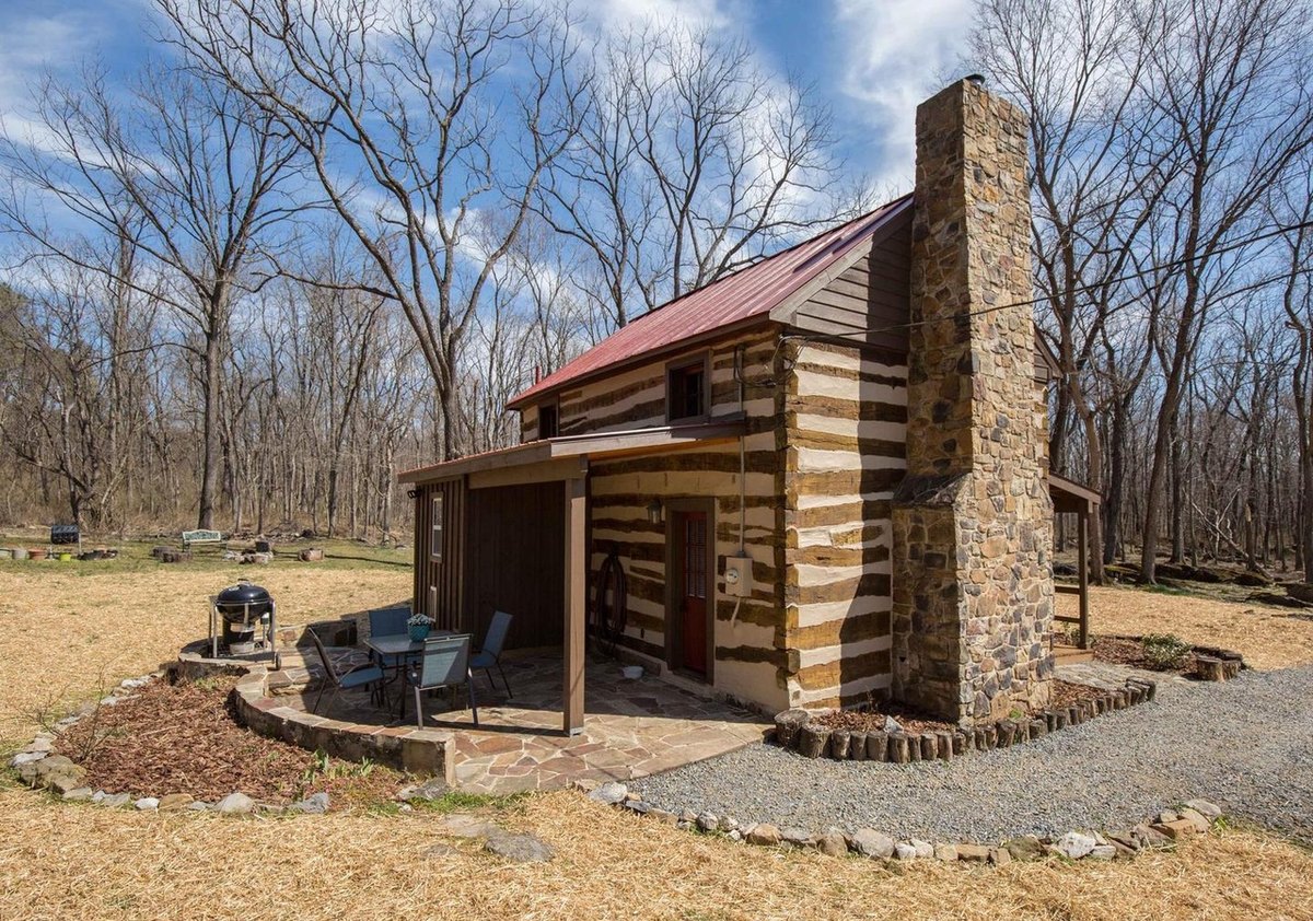 The back corner of the cabin with the fieldstone chimney, a covered flagstone patio with a table and chairs, and a grill on a curved stone wall.