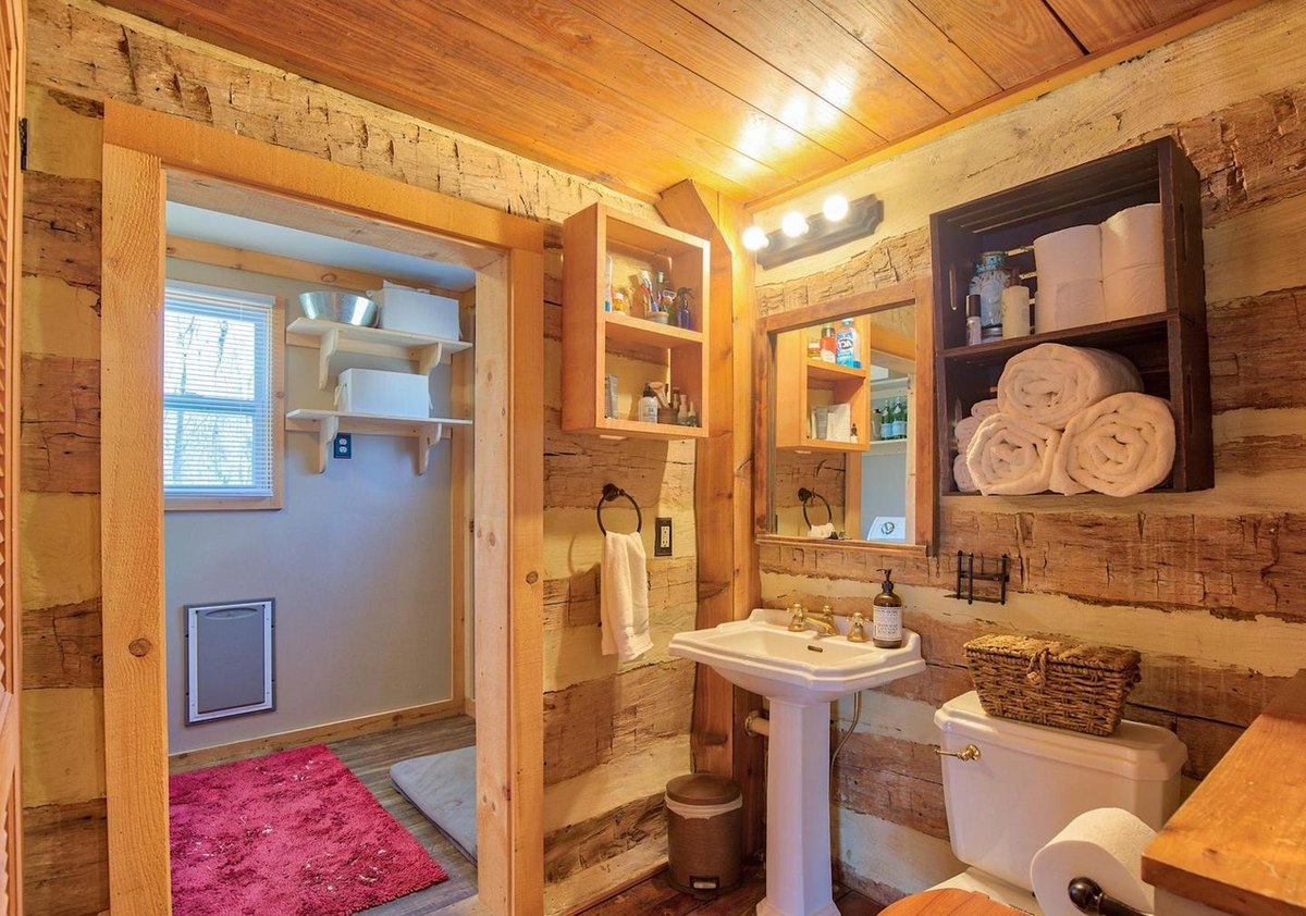 The bathroom with log walls: a pedestal sink under a mirror and lights, open shelves with rolled towels, and a doorway through to the laundry room.