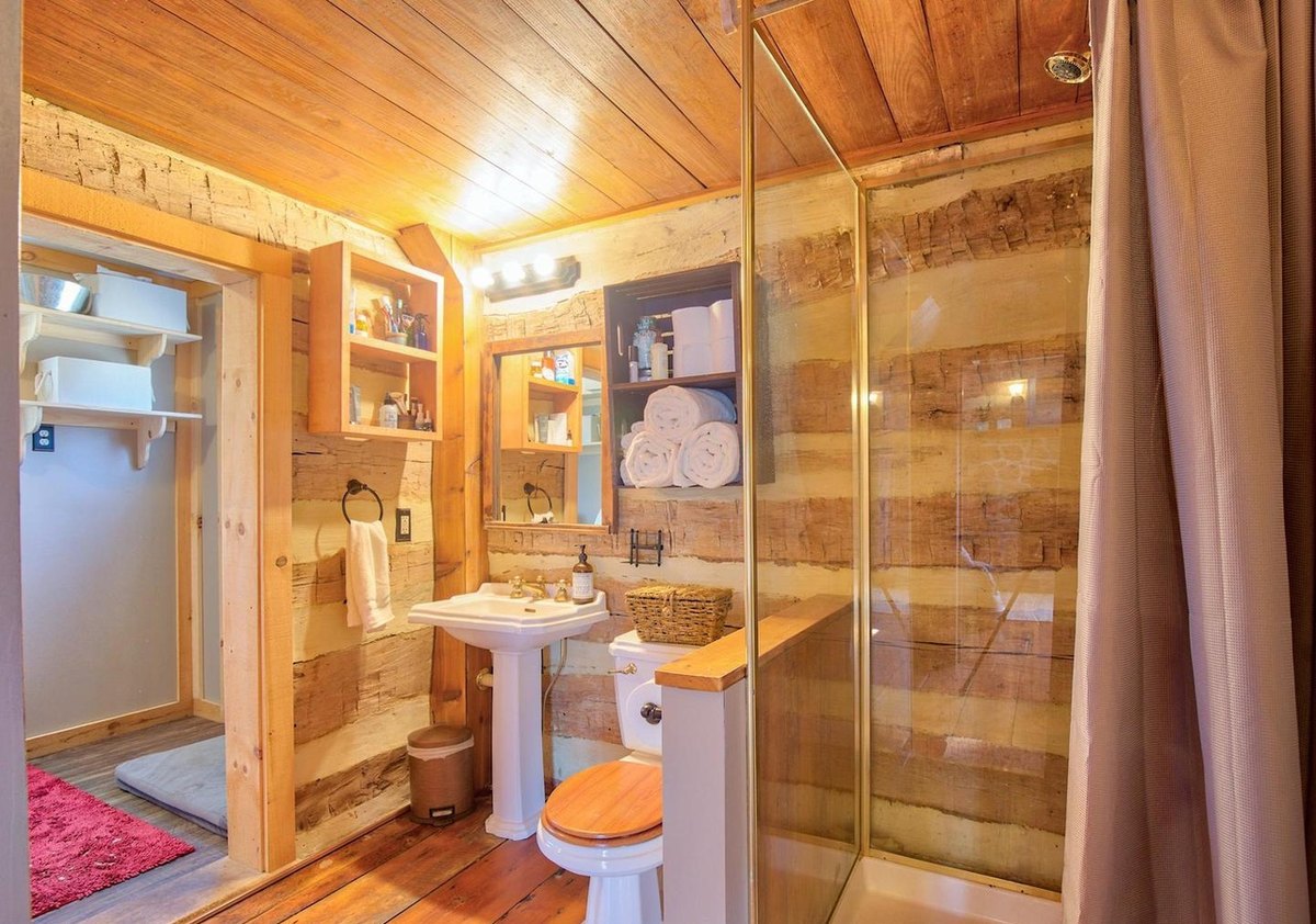 The bathroom showing the pedestal sink and shelves beside a glass shower enclosure and a curtain, with log walls and a board ceiling.