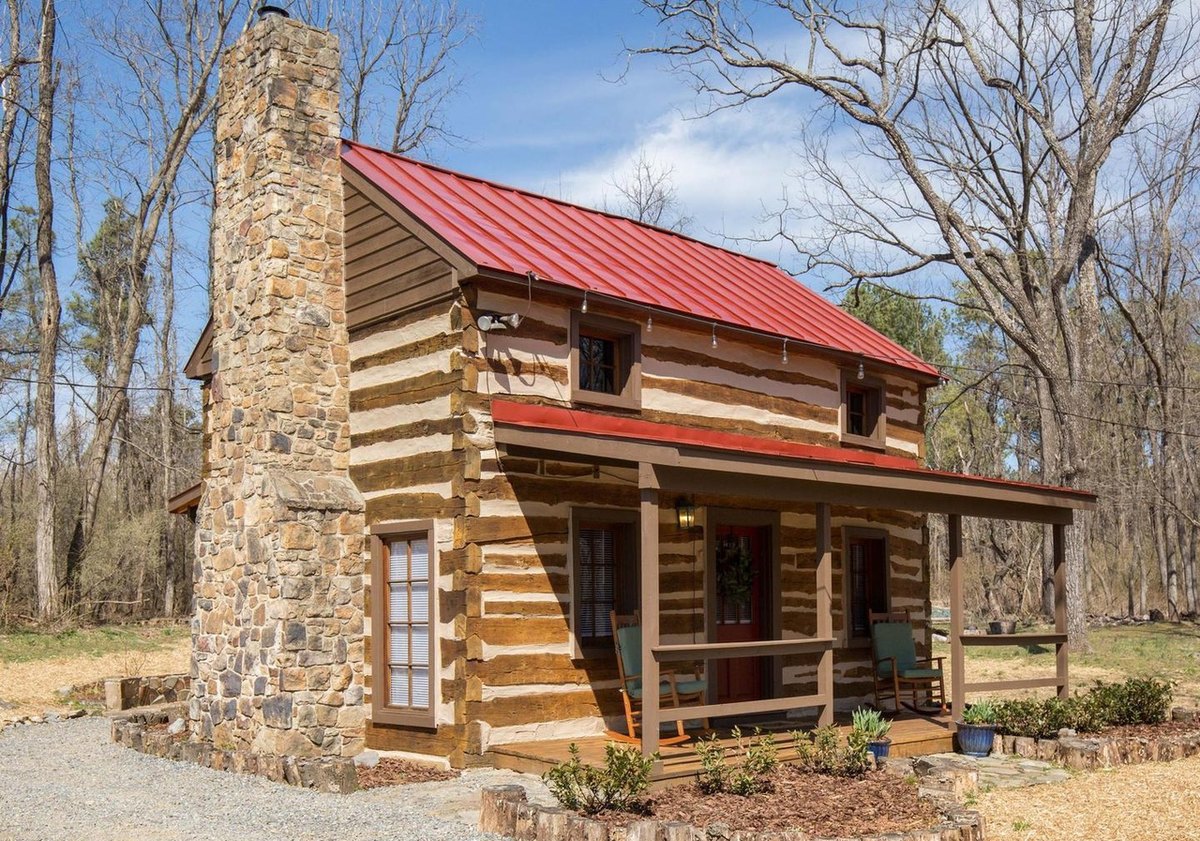 The cabin from the chimney-end corner: a tall fieldstone chimney, a glazed door, the red roof and the covered front porch with green chairs, and a gravel path around it.