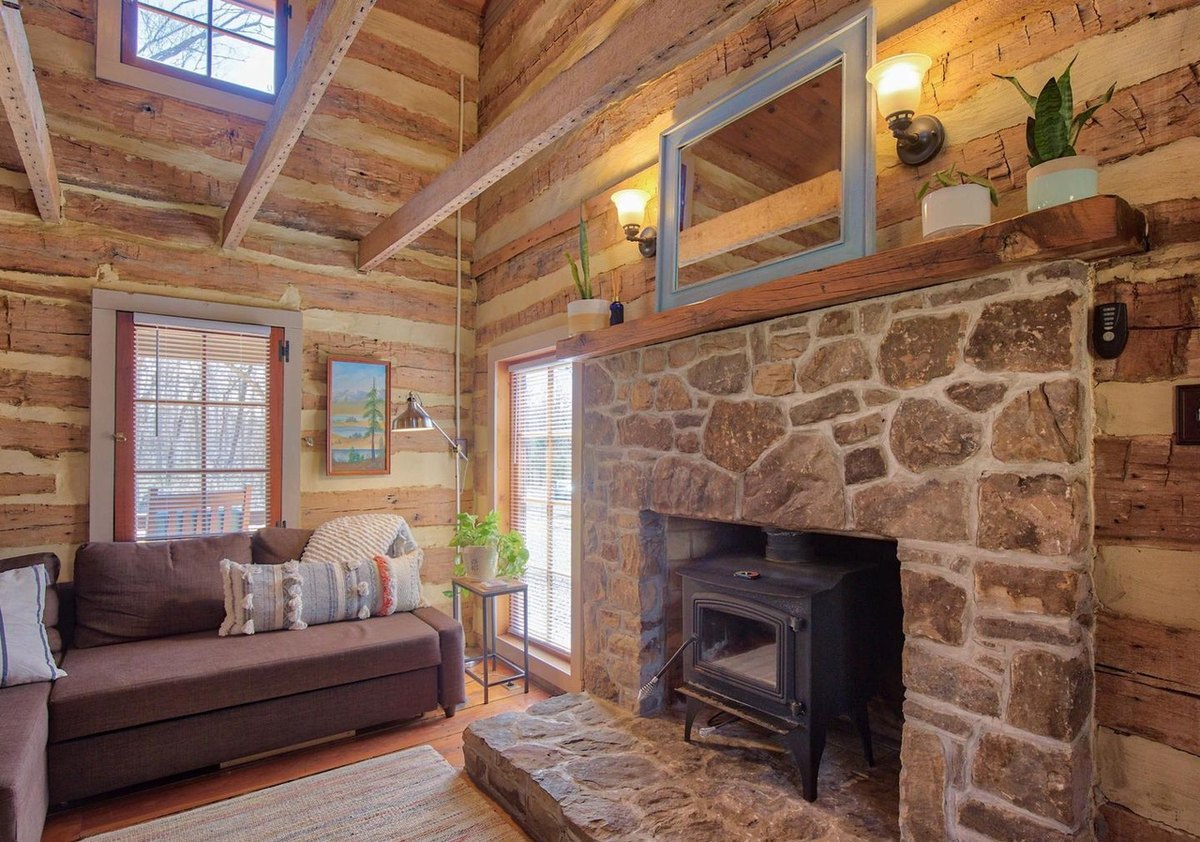 The stone fireplace with a black wood stove on its raised stone hearth, a mirror and plants on the wooden mantel, and a sofa beside a window.