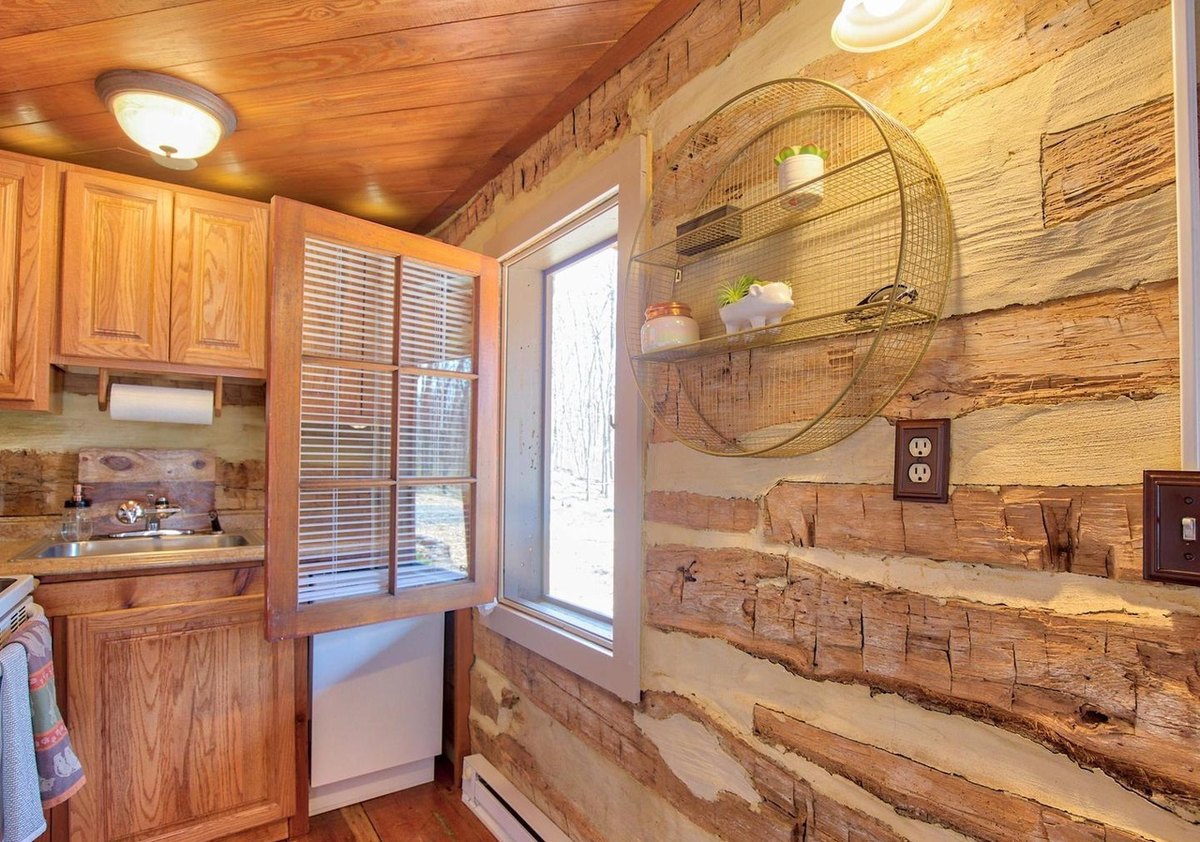 The kitchen from its doorway, showing a wall of broad hewn logs with pale chinking, a window with blinds, and cabinets and a sink at the left.