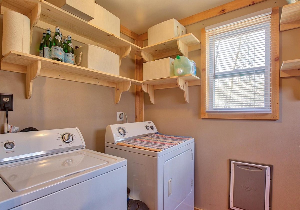 A small laundry room with a washer and dryer under wooden shelves, a window with blinds and pale painted walls.