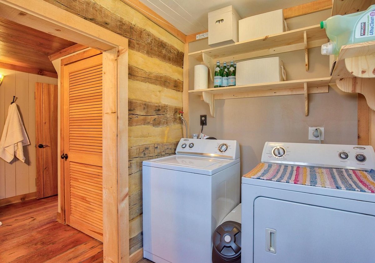 The laundry room from its doorway, with the washer and dryer, shelves above, a log wall at the left and a louvered door.
